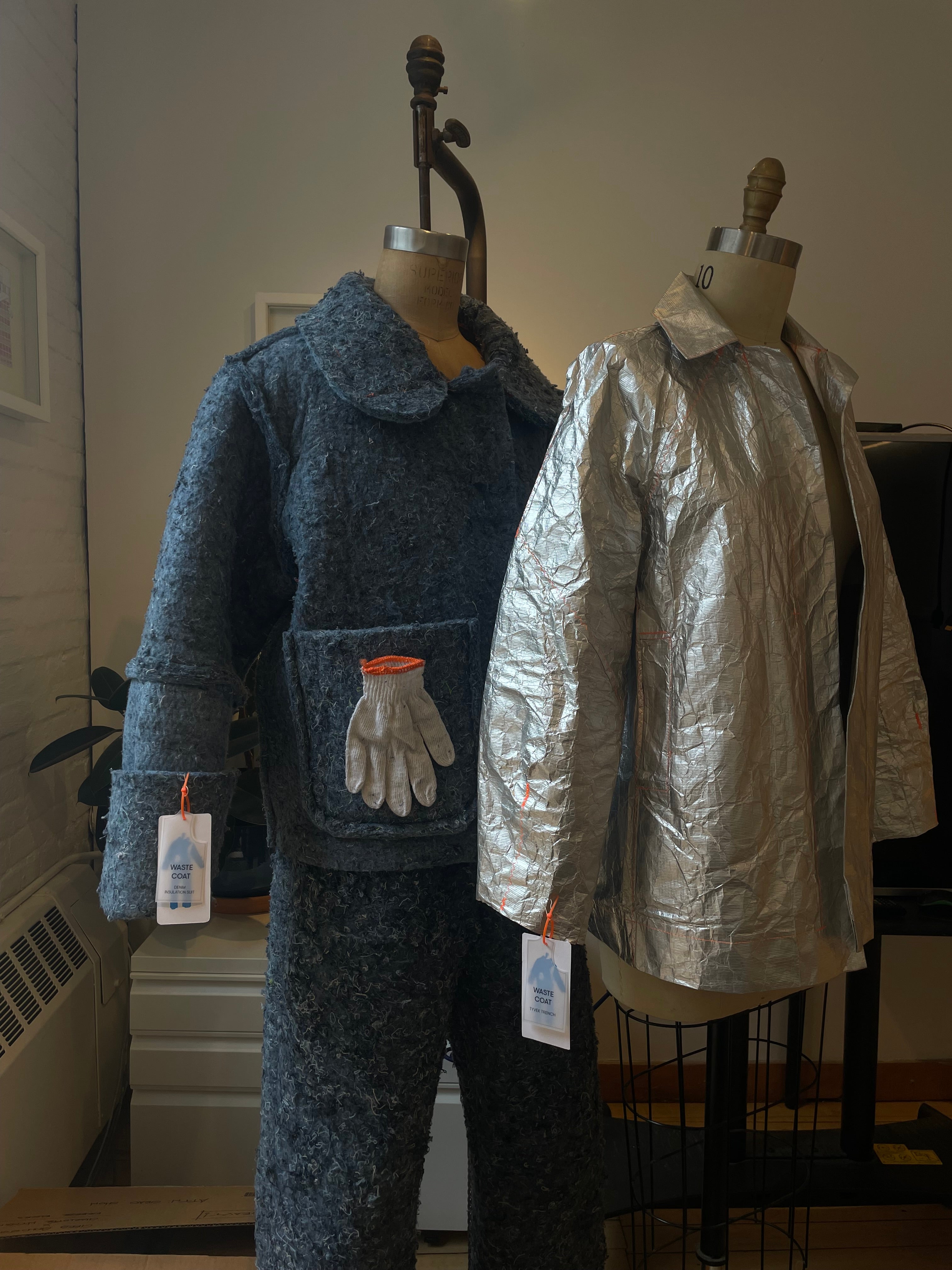 /Two%20jackets%2C%20one%20made%20from%20recycled%20denim%20insulation%2C%20the%20other%20from%20silver%20Tyvek%2C%20on%20two%20mannequins%20with%20garment%20tags%20that%20say%20%22Waste%20Coats%22