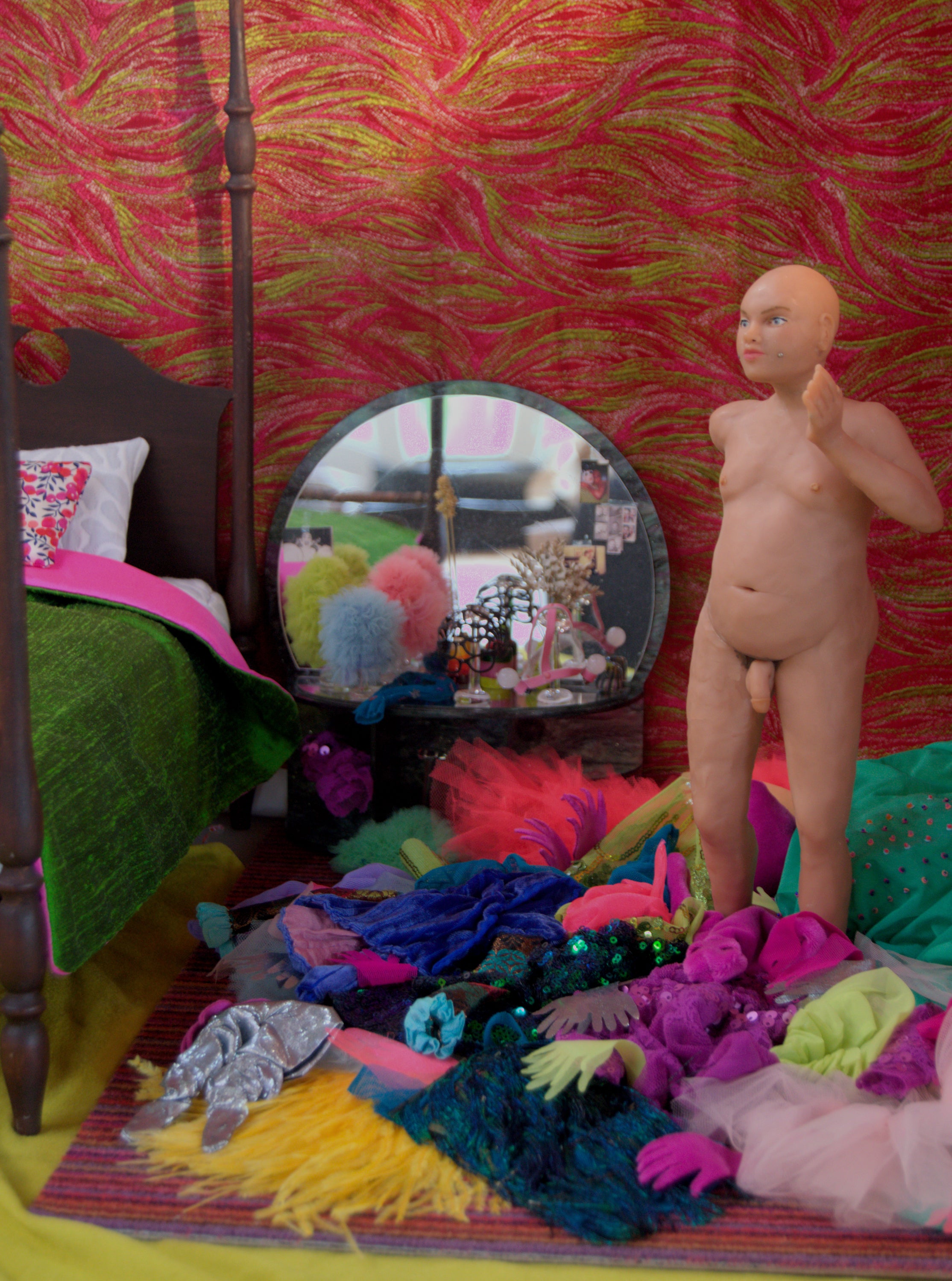 /A%20sculpted%20nude%20figure%20stands%20in%20a%20bedroom%2C%20the%20floor%20covered%20with%20elaborate%2C%20brightly-colored%20clothes.%20A%20mirrored%20vanity%20has%20ruffled%20headdresses%20on%20glass%20wigstands%2C%20a%20post%20bed%20is%20covered%20with%20a%20green%20and%20pink%20silk%20duvet.