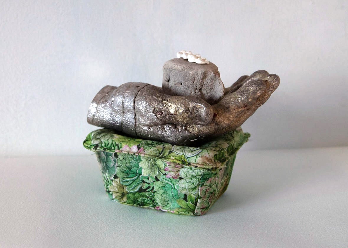 /a%20stack%20of%20objects%3B%20a%20white%20mold%20of%20three%20molar%20teeth%2C%20an%20ocean%20animal%20vertebrae%2C%20a%20pewter%20hand%20that%20is%20crushing%20a%20blackberry%20plastic%20container%20that%20is%20covered%20in%20green%20leaf%20patterned%20fabric