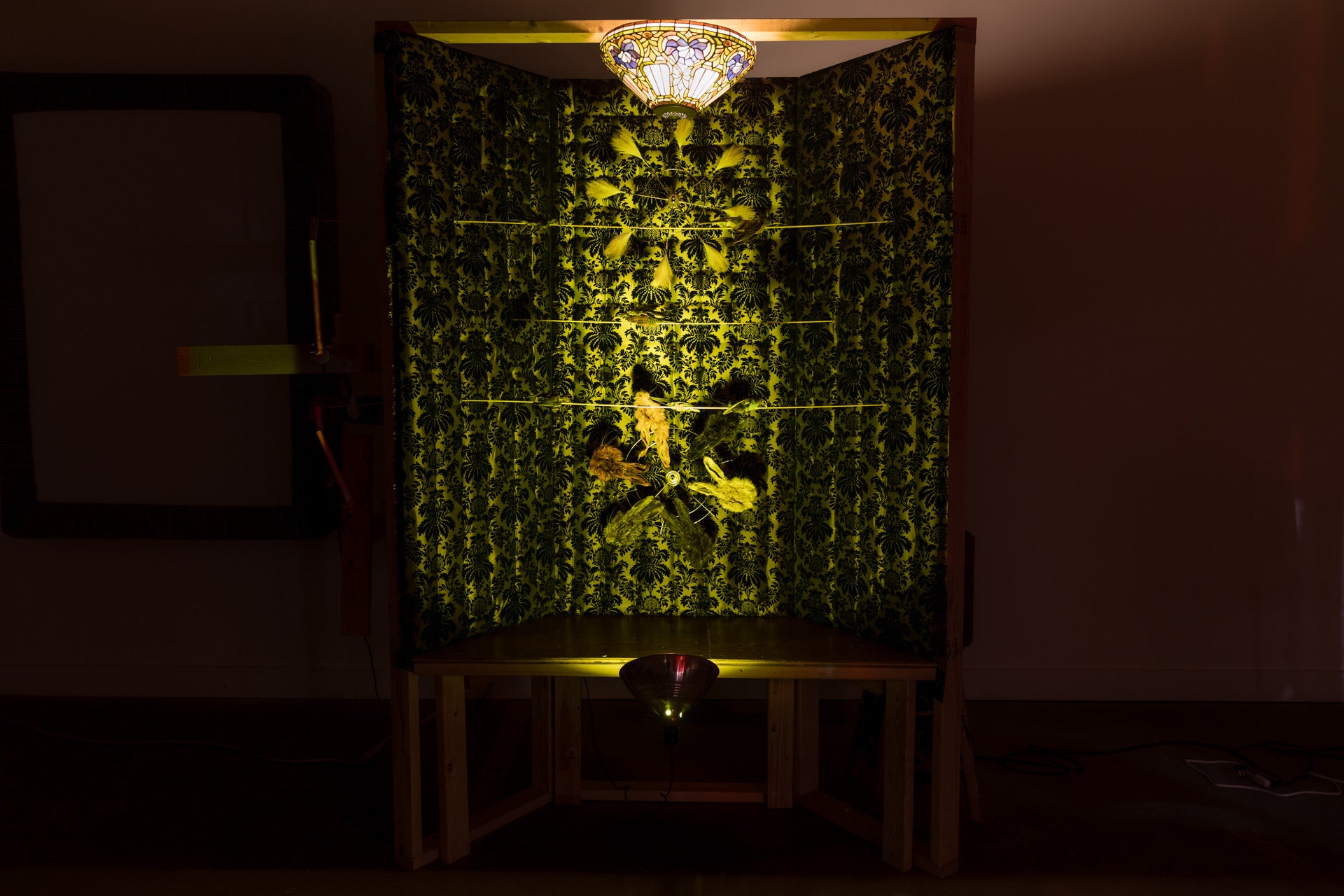 /A%20fabric%20covered%203%20paneled%20wall%20the%20moving%20rabbit%20maks%2C%20and%20spinning%20taxidermy%20birds%20moving%20throughout.%20A%20faux%20tiffany%20lamp%20emits%20a%20yellow%20light.