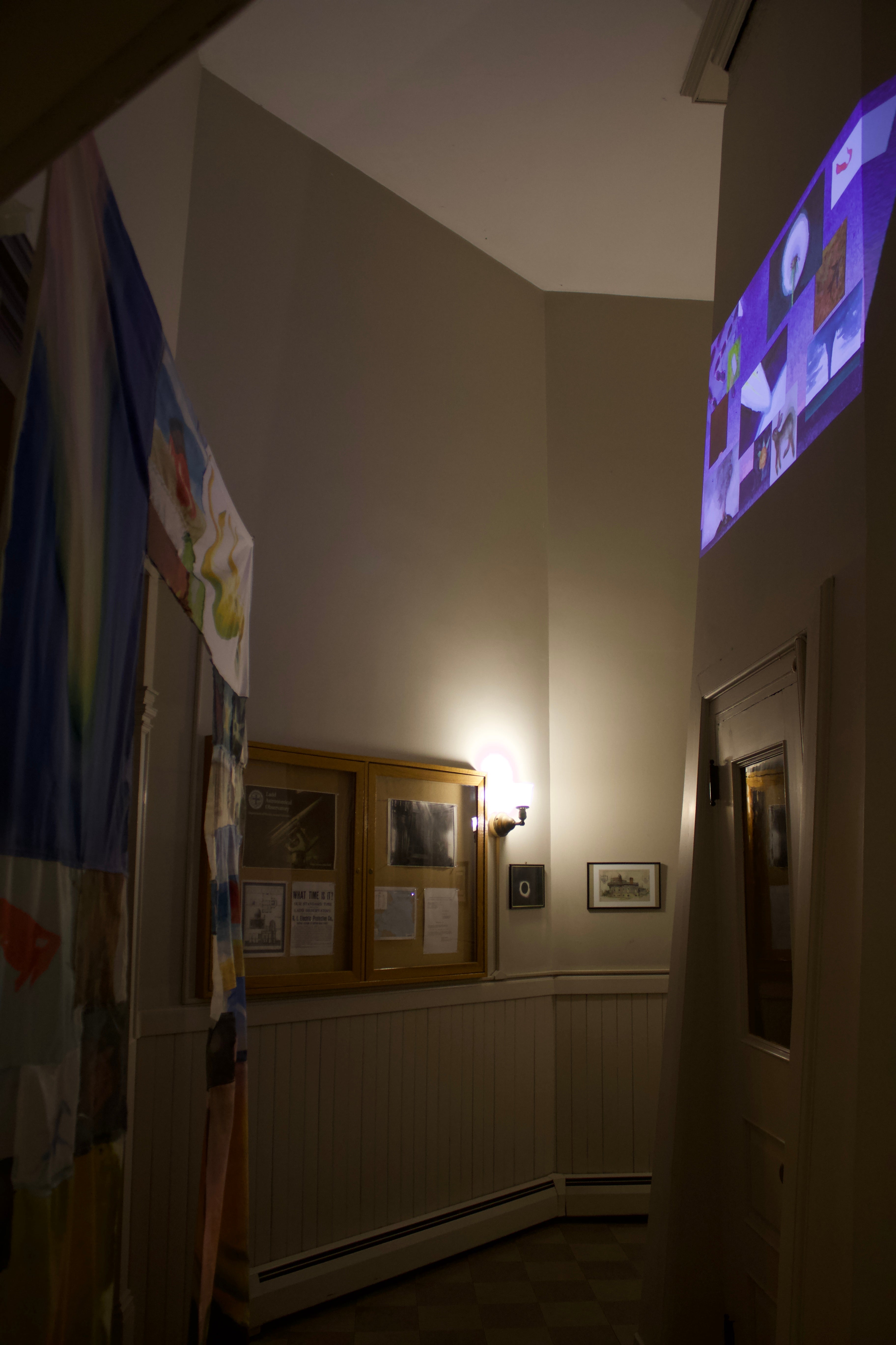 /An%20image%20of%20a%20darkened%20interior%20space%2C%20a%20doorway%20covered%20in%20fabric%20and%20an%20animation%20with%20multiple%20moving%20windows%20projected%20on%20opposite%20wall