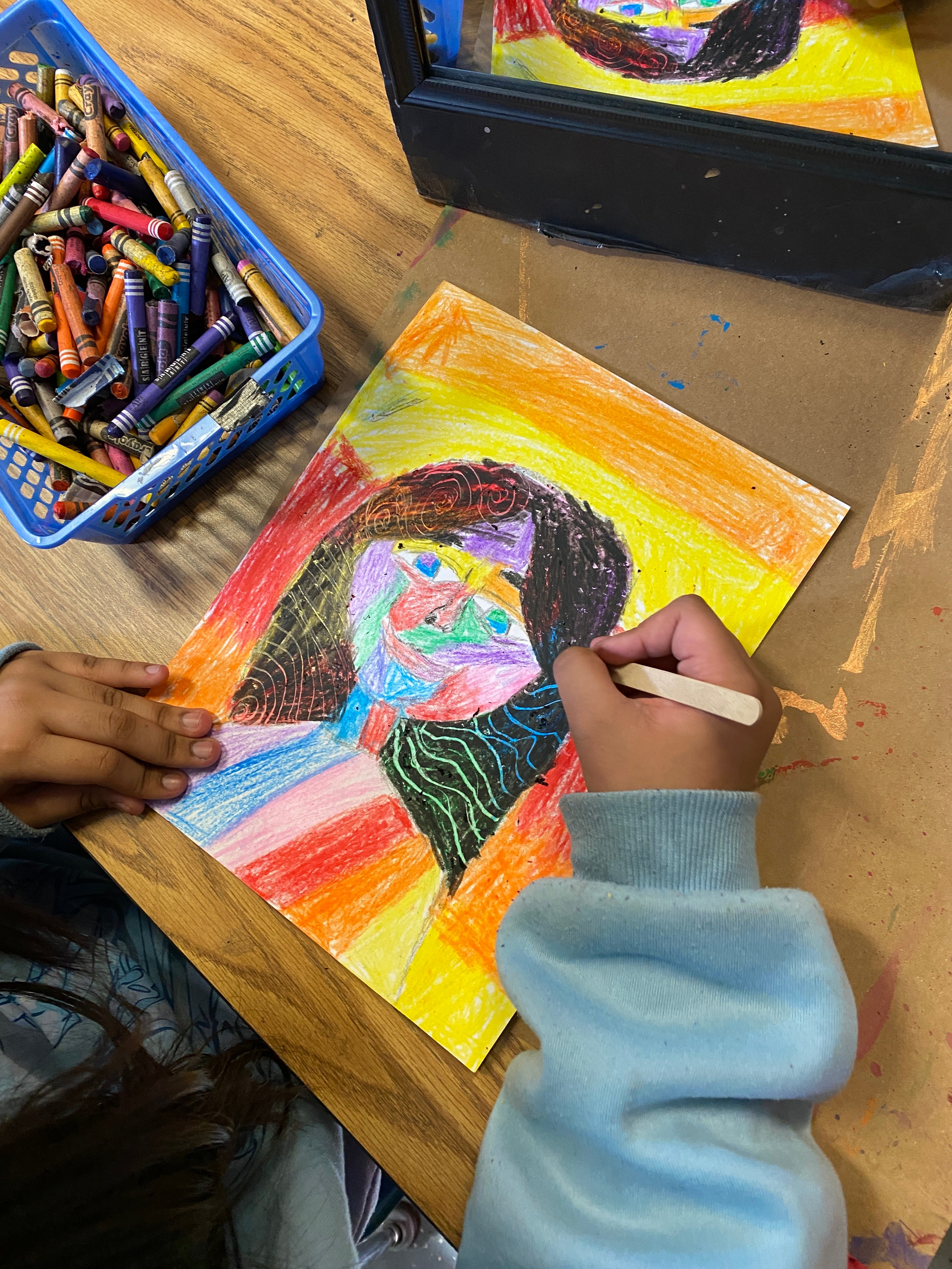 A student's hand holding a popsicles stick is scratching into a crayon-laden paper. The popsicle stick is creating patterns in the self portrait. A bin of crayons can be seen to the left of the image.