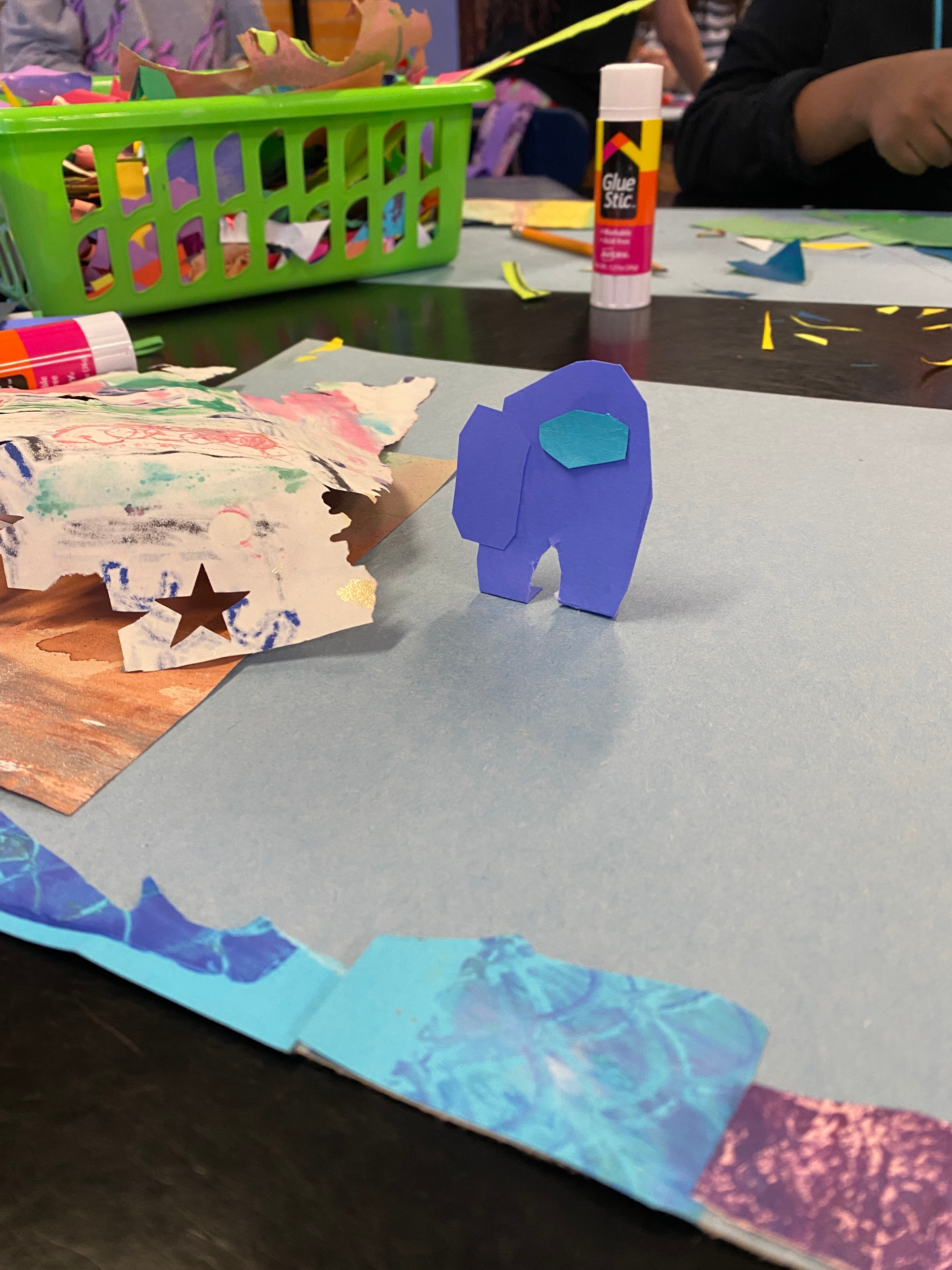 A paper collage cut out of a spaceman from the video game Among Us stands up in a 3D fashion on a student's paper. Seen in the background is a mess of papers and gluesticks.
