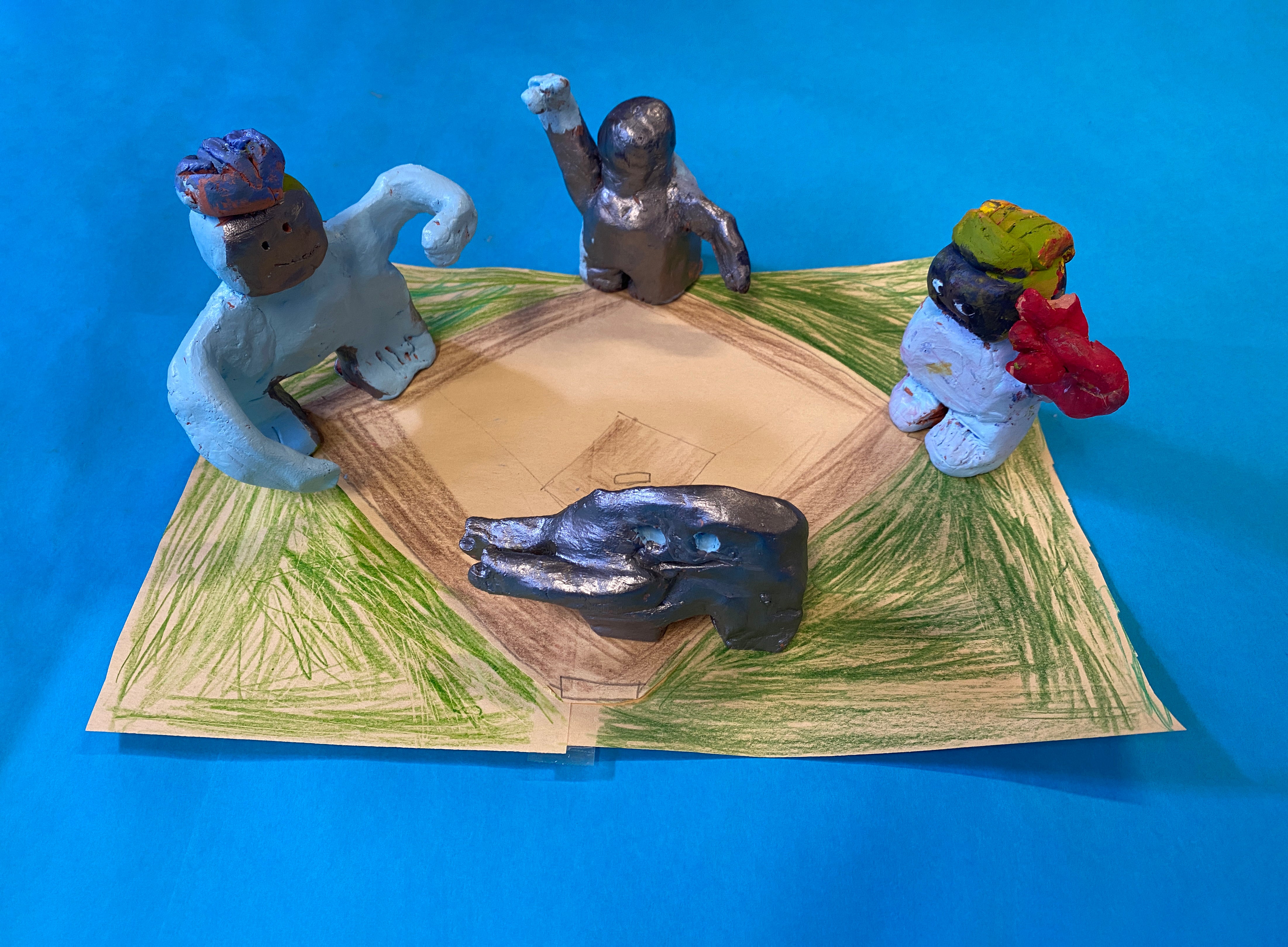 Four clay figurines stand on a hand drawn baseball diamond atop a bright blue backdrop. The figurines have been painted a combonation of silver, light blue, and red, and all have some version of outstretched arms.
