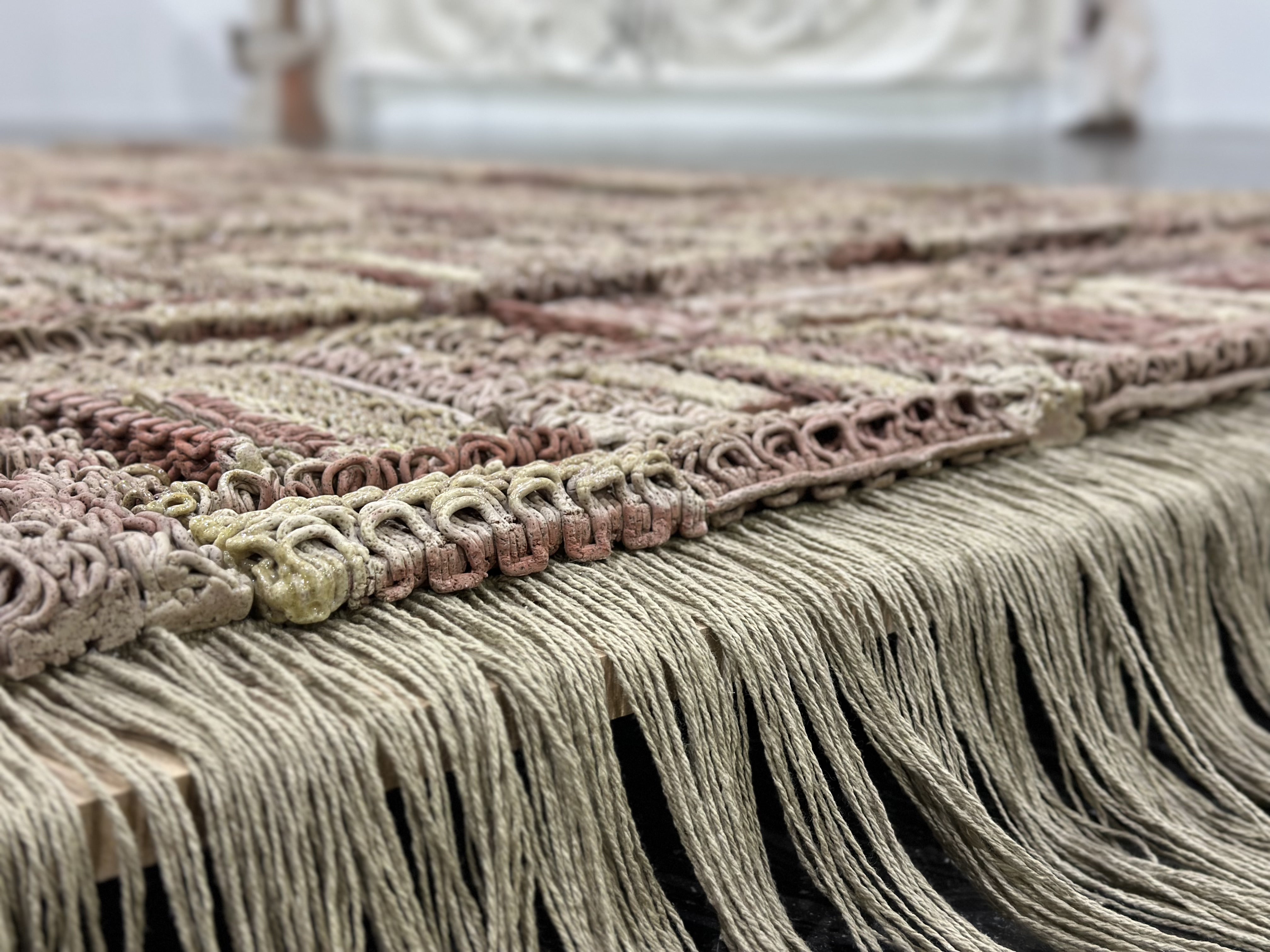 3D-printed ceramic (Egyptian paste) piece with intricate patterns in shades of red, beige, and brown. The first row is made of yarn with hanging threads. The piece is displayed on a wooden platform, placed on a polished concrete floor.