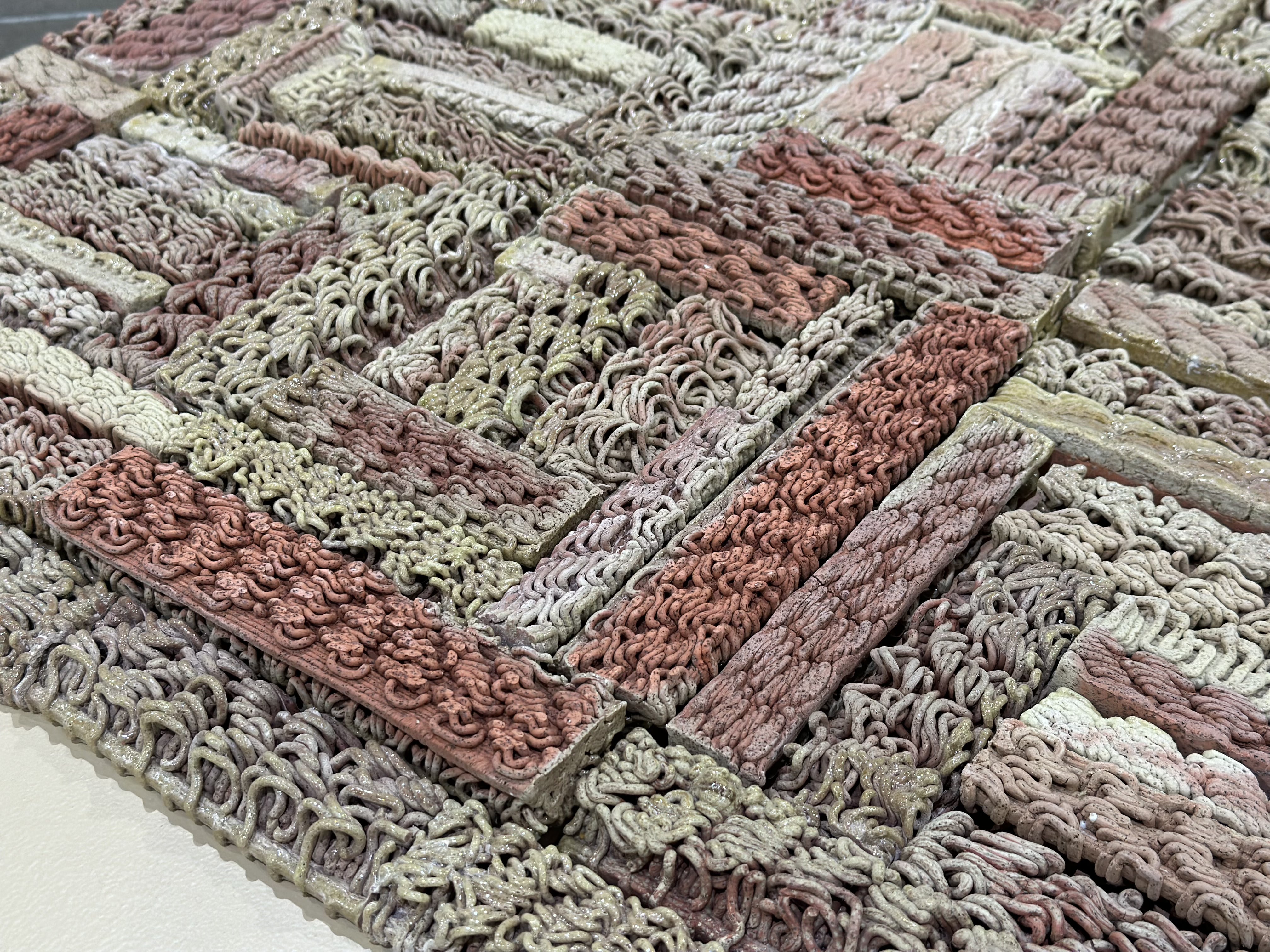 3D-printed ceramic (Egyptian paste) piece with intricate patterns in shades of red, beige, and brown. The first row is made of yarn with hanging threads. The piece is displayed on a wooden platform, placed on a polished concrete floor.