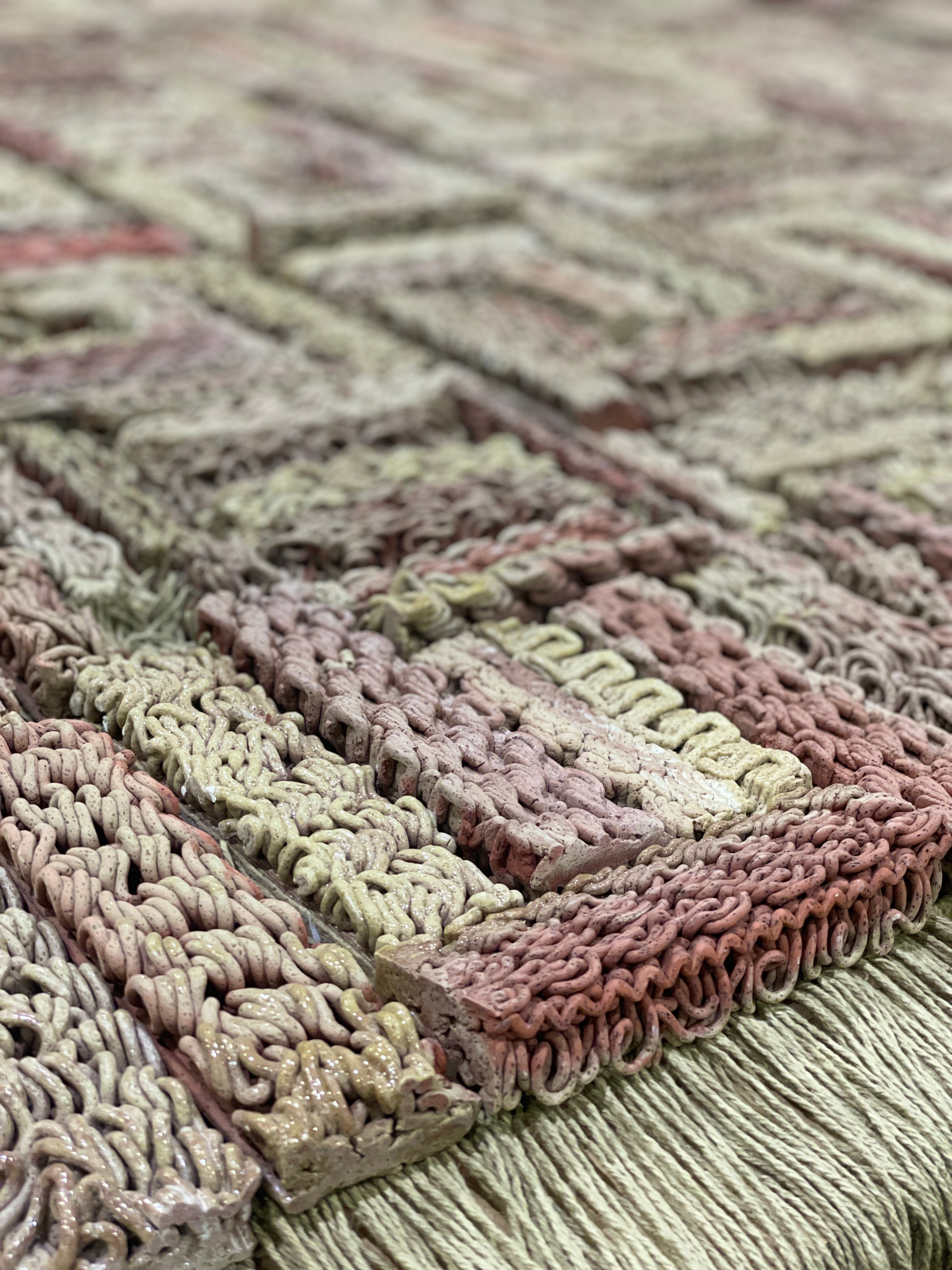 3D-printed ceramic (Egyptian paste) piece with intricate patterns in shades of red, beige, and brown. The first row is made of yarn with hanging threads. The piece is displayed on a wooden platform, placed on a polished concrete floor.