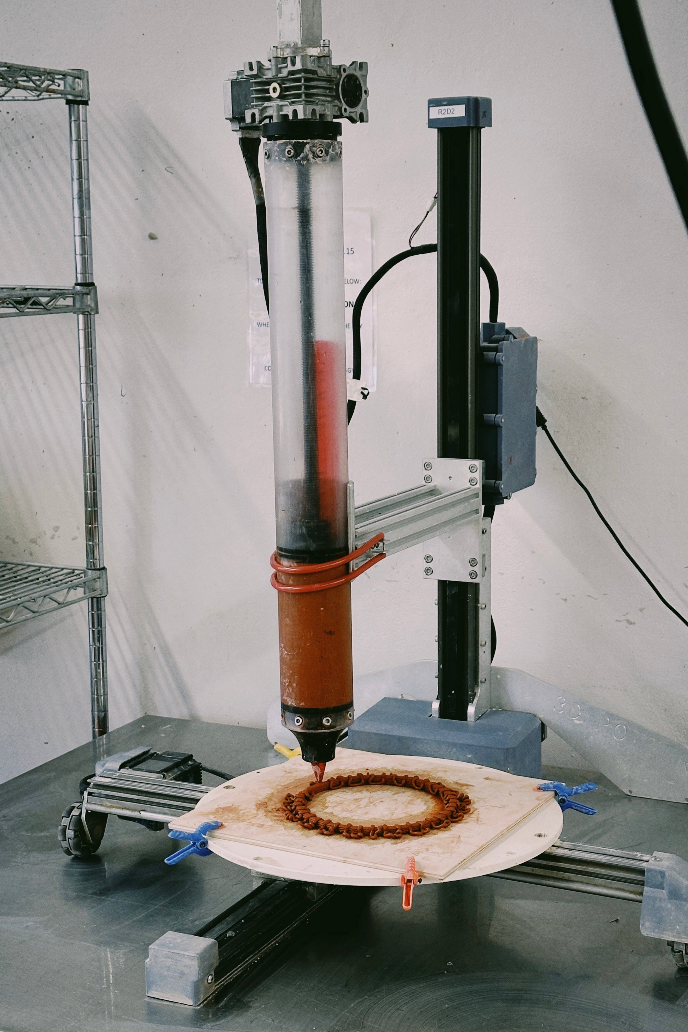 /3D%20printer%20extruding%20a%20circular%20clay%20pattern%20on%20a%20rotating%20platform%2C%20with%20the%20printer%20labeled%20%27R2D2%27%20in%20a%20workshop%20setting.