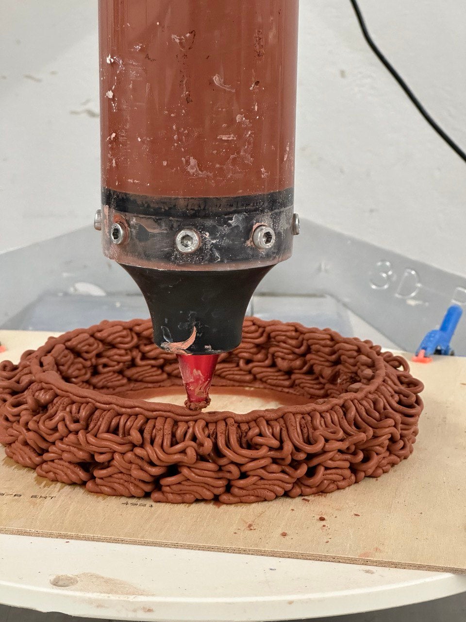 /3D%20printer%20nozzle%20depositing%20intricate%2C%20layered%20clay%20structure%20onto%20a%20wooden%20platform%2C%20capturing%20the%20detailed%20formation%20process.