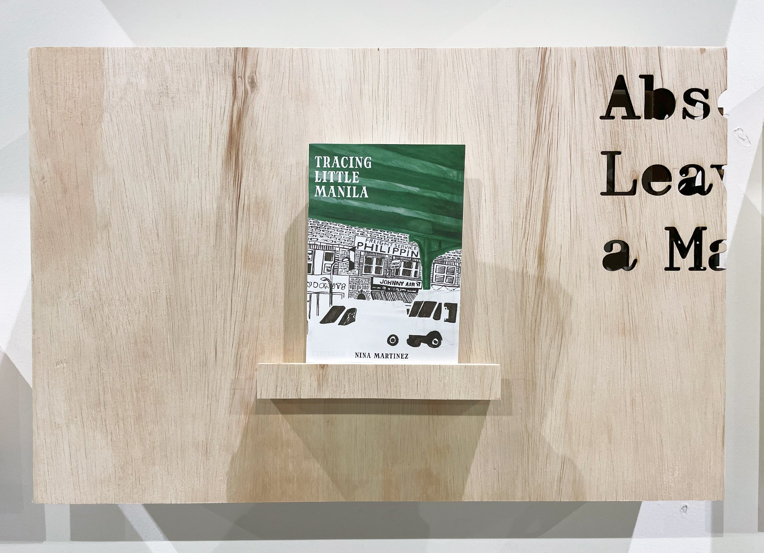 A closeup of the Tracing Little Manila zine, sitting on a small shelf on a plywood display. The text "Absence Leaves a Mark," the title of my thesis, is laser cut into the corner.