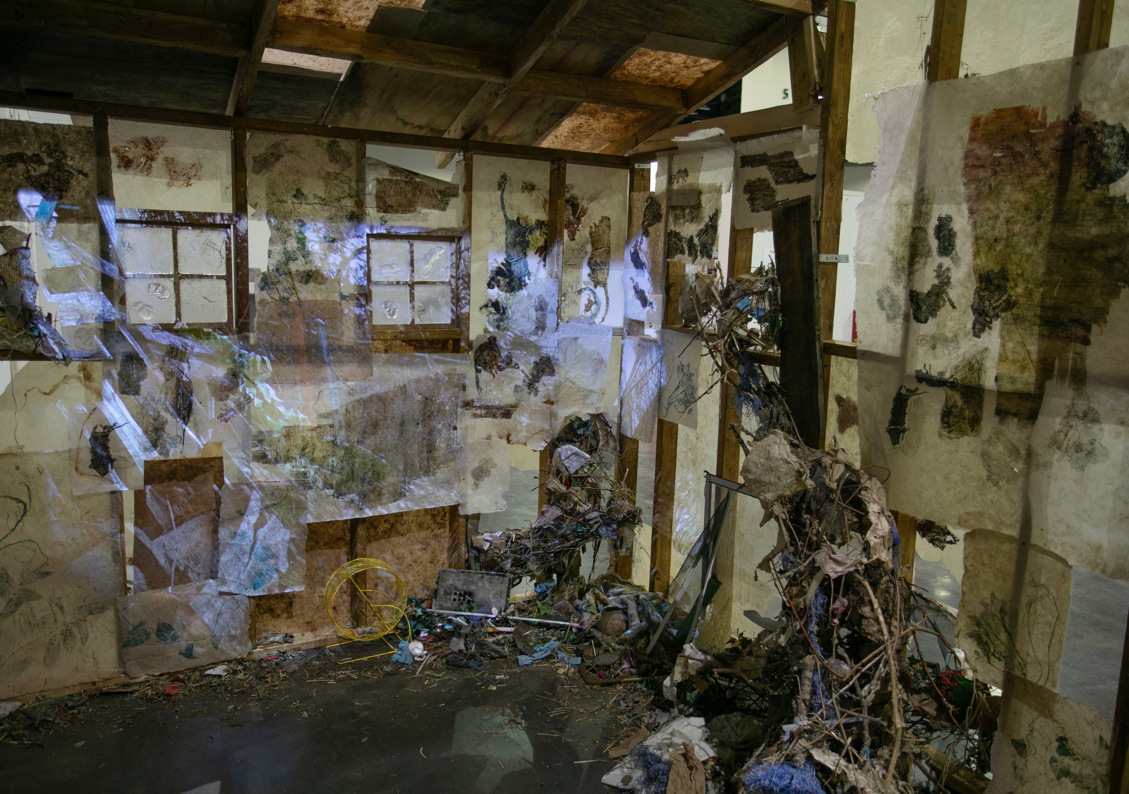 Inside a structure with exposed framing, two windows and print-lined walls, with nest-like forms going into the walls, and a projection of a pile of a pile of wood in a garden