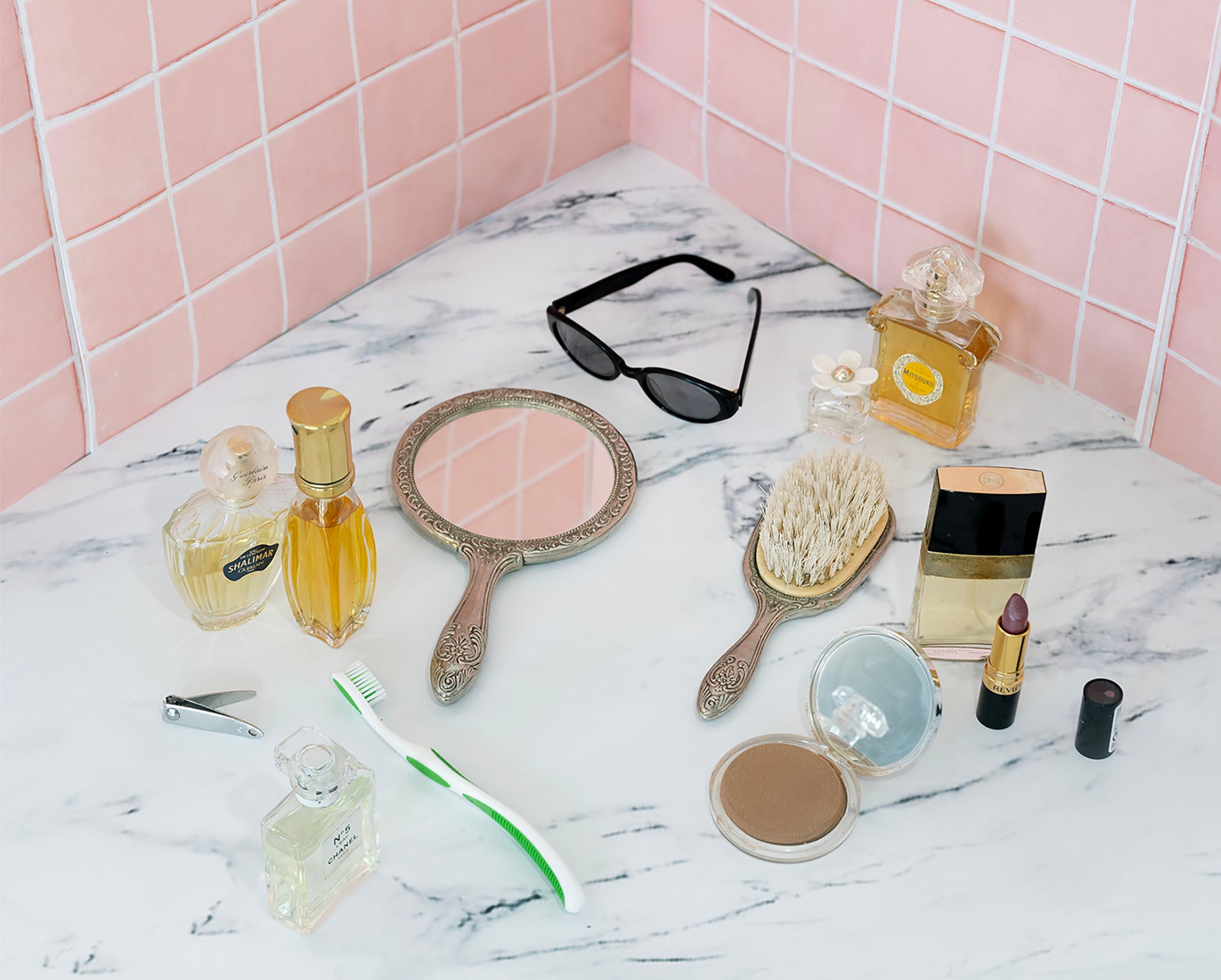 /A%20still%20life%20of%20a%20marble%20countertop%20against%20pink%20ceramic%20tiles%20with%20perfume%20bottles%2C%20a%20hairbrush%2C%20compact%20mirror%2C%20sunglasses%2C%20and%20makeup.