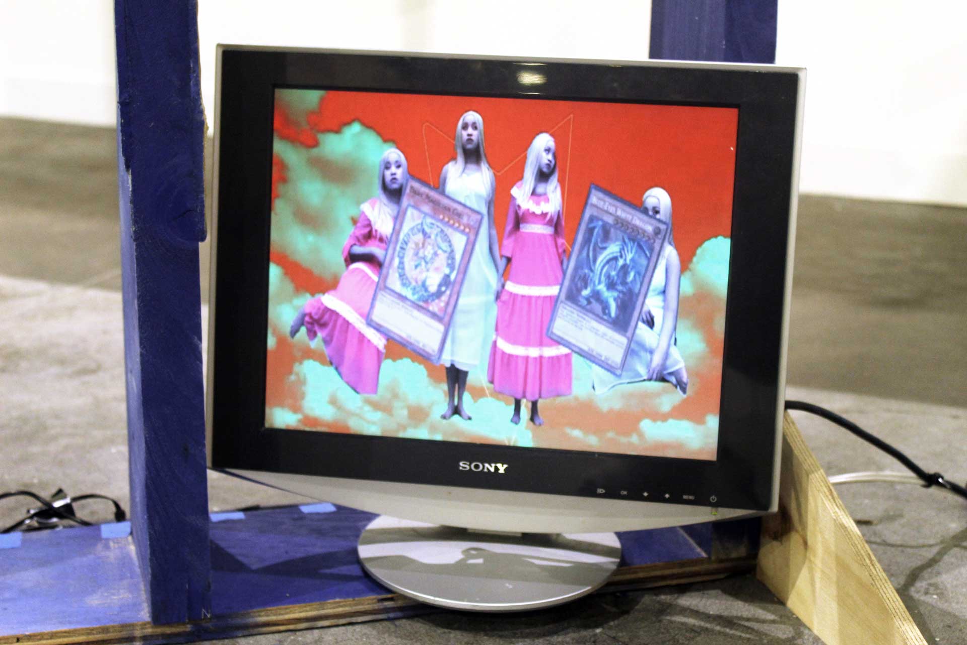 A still from the video on a silver Dell monitor shows the artist four times in pale makeup, wearing a bright pink period dress and a pale blue nightgown, symmetrically on a red and green horizon. A pentagram rotates in the background.