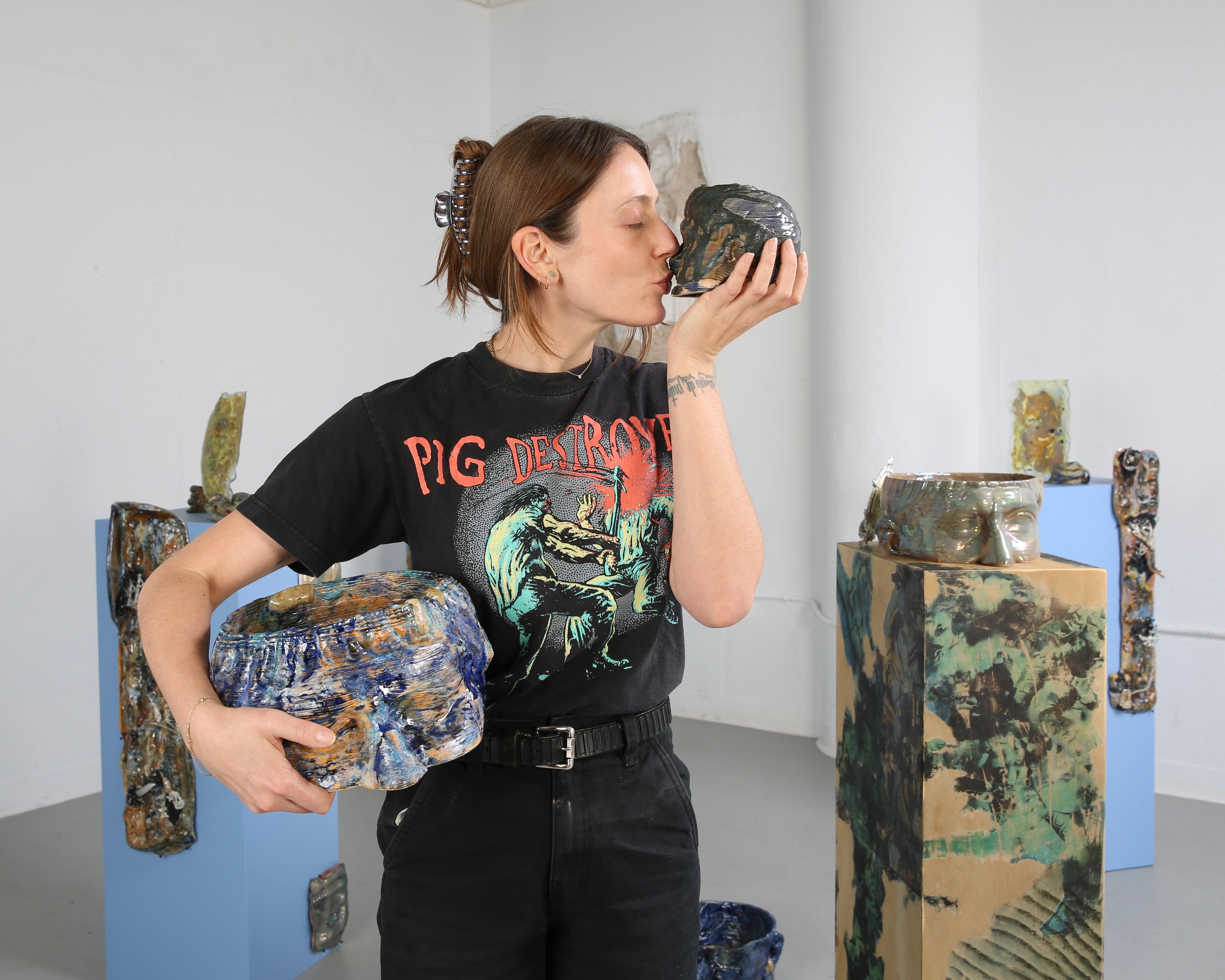 Portrait of artist standing in front of her installation. Turned to her left and kissing a small ceramic 3D printed head.   