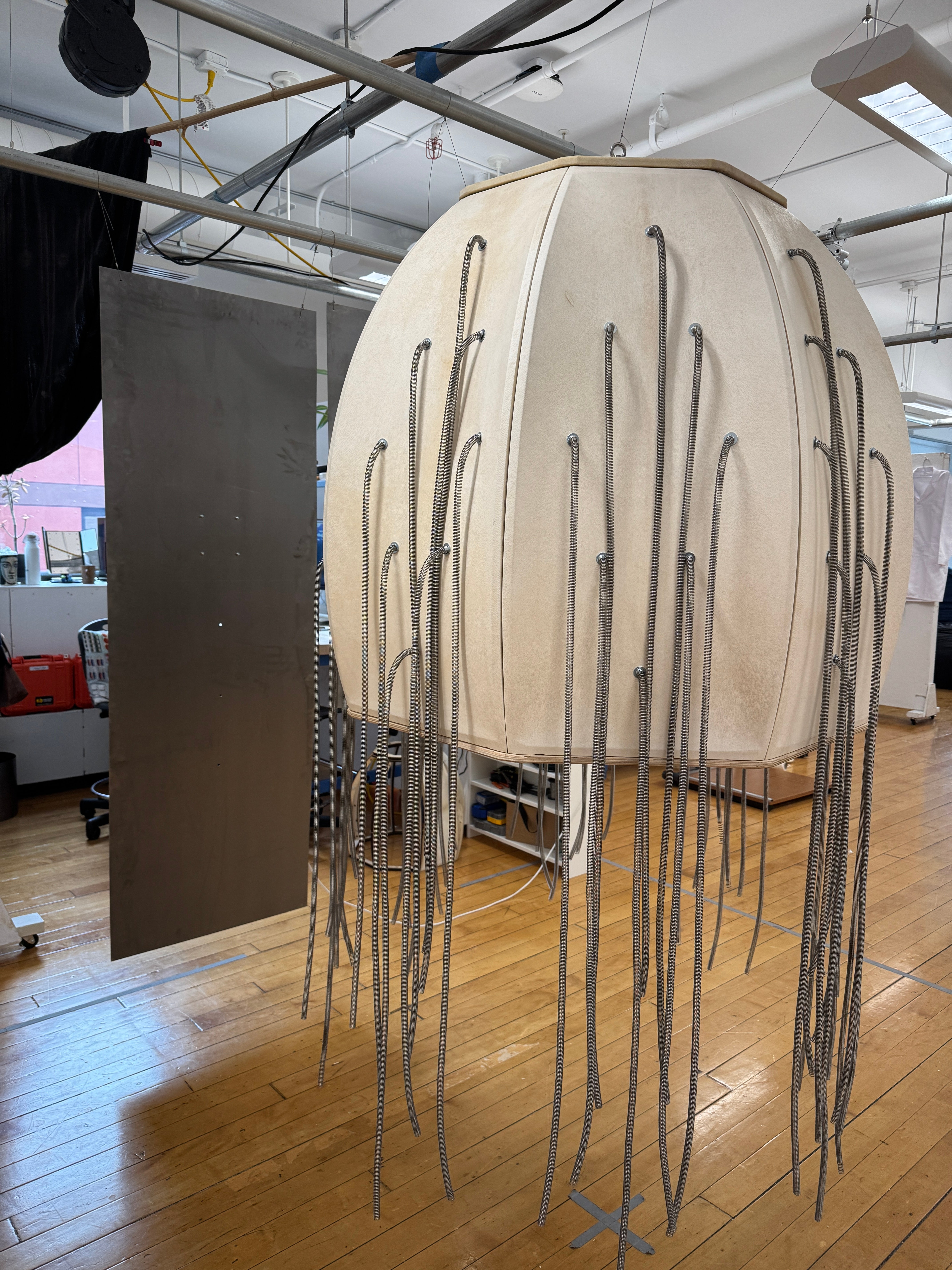 Barrel-shaped goatskin structure with hanging metal springs, surrounded by steel sheets and lit by overhead projection.