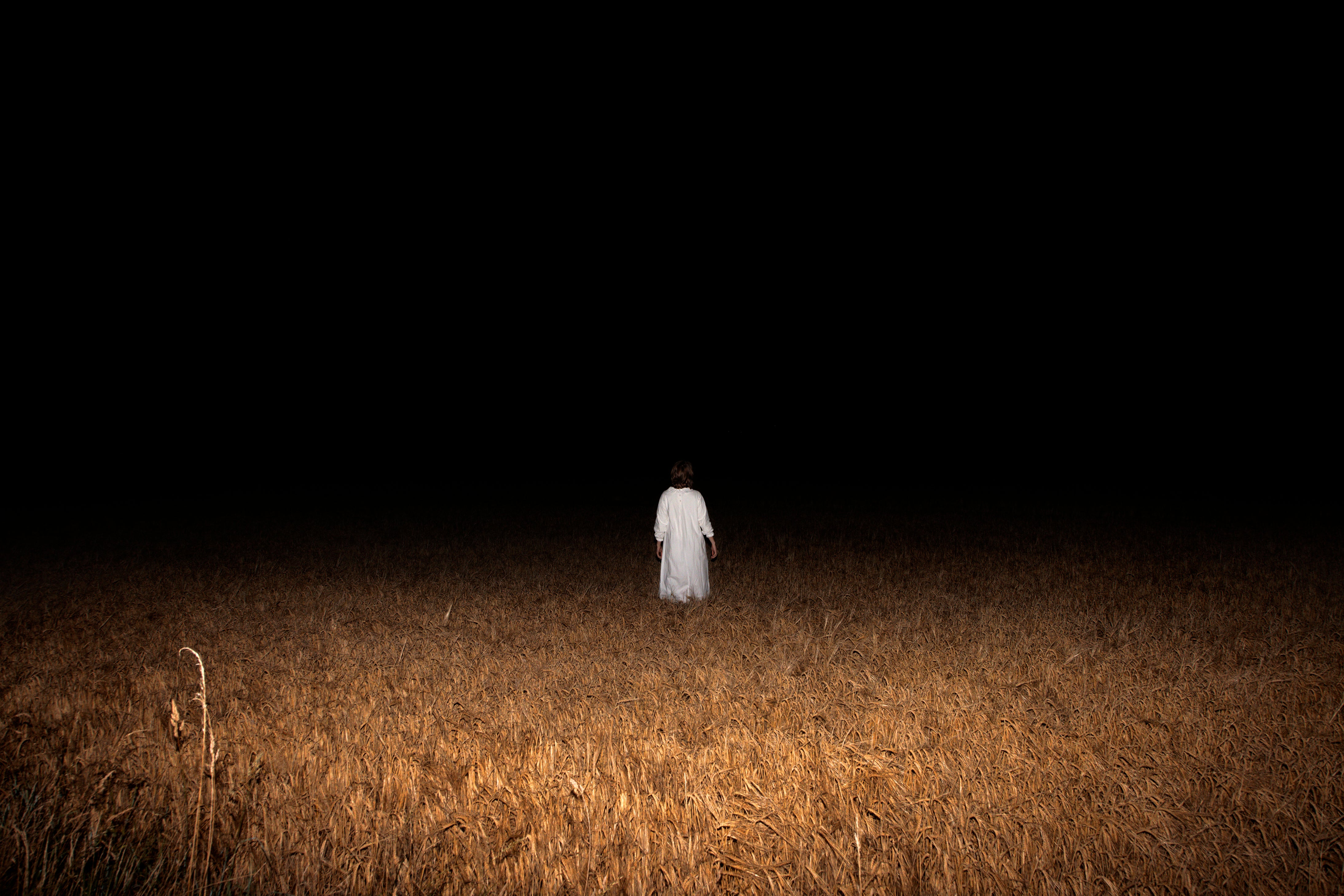 A figure in a white Sankta Lucia gown stands in a wheat field. She faces the dark, void of the night. 