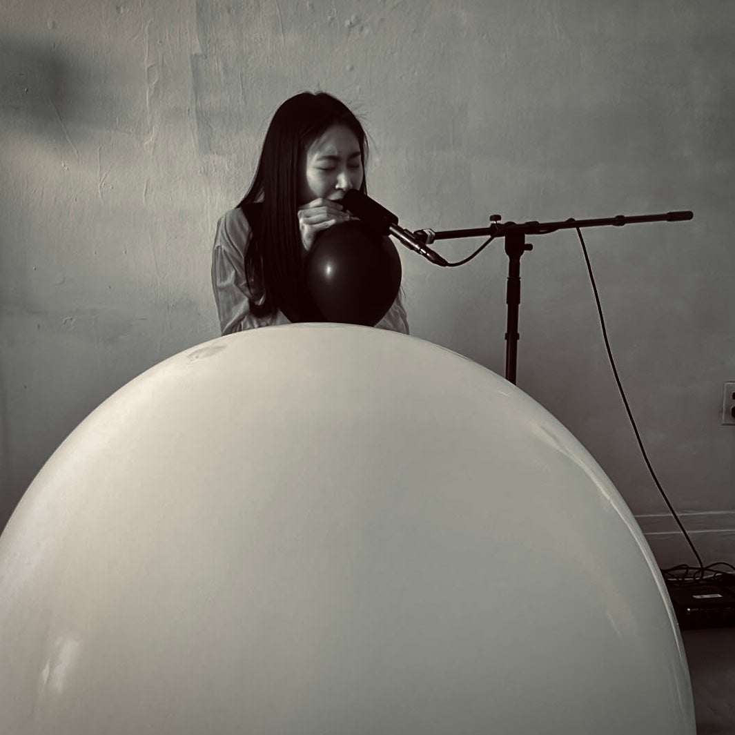 A performer creates balloon noise with a microphone, processed through a computer, and played back via speakers.