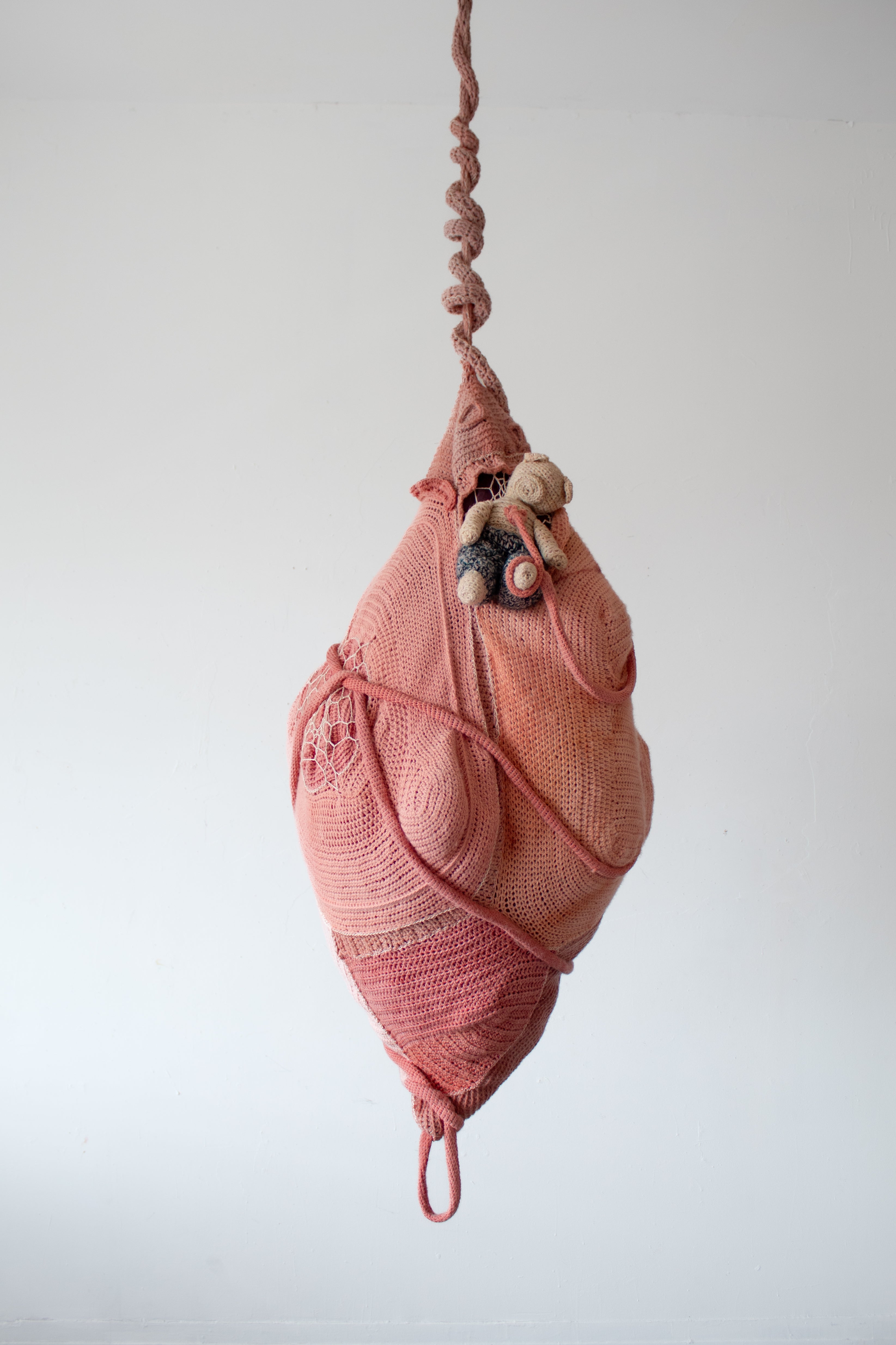 pink, pupa-shaped crochet sculpture with tapering ends, hanging by a twisted cord; a small crochet teddy bear emerges.