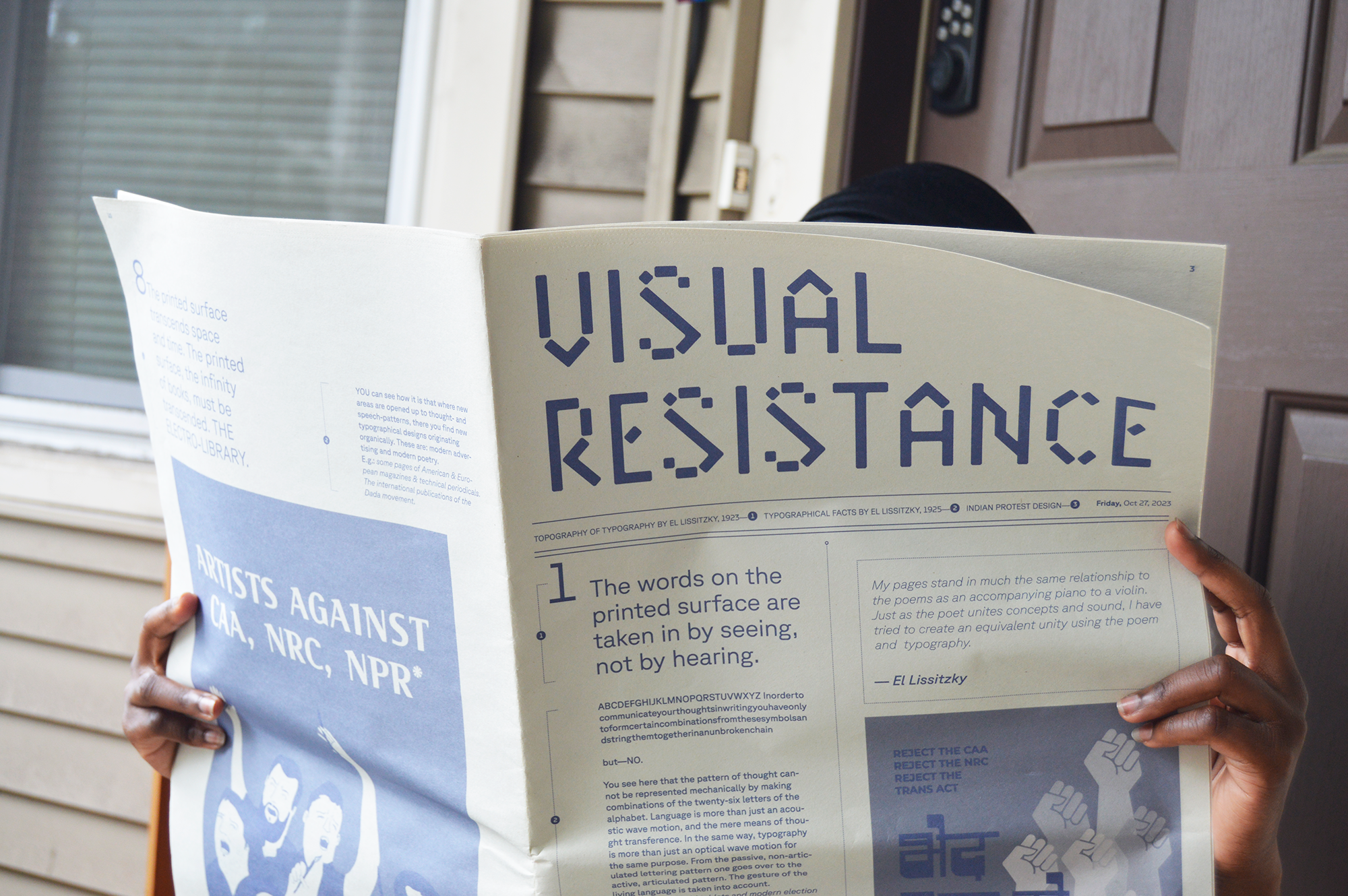 Person holding open 'Visual Resistance' newspaper, face hidden, hands gripping the spread.