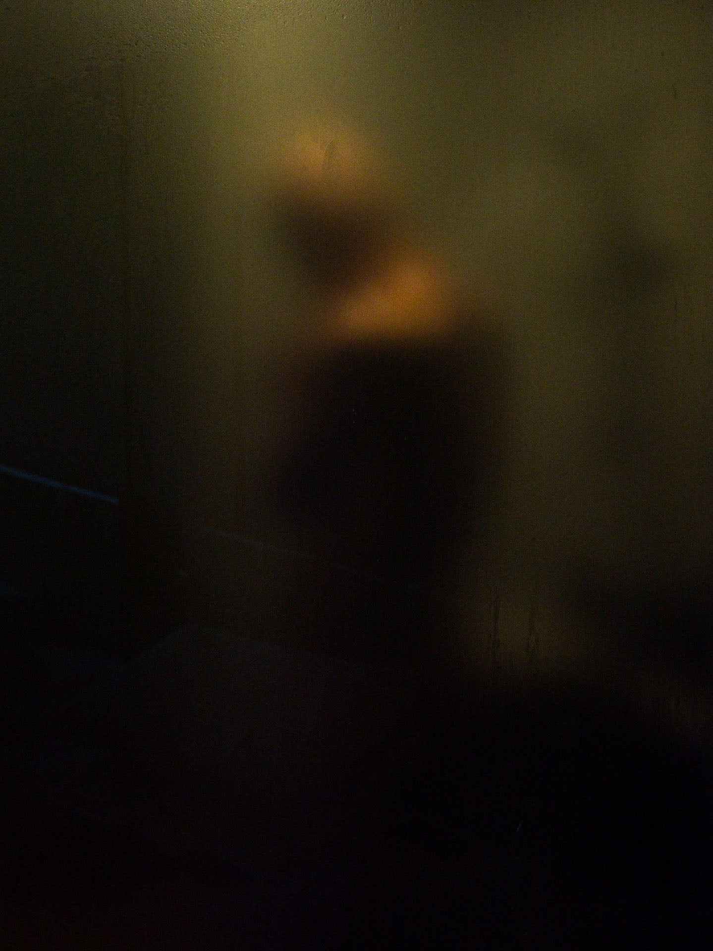 A shadowy silhouette of a man bathing behind translucent hotel glass, soft light creating intimacy and emotional distance.
