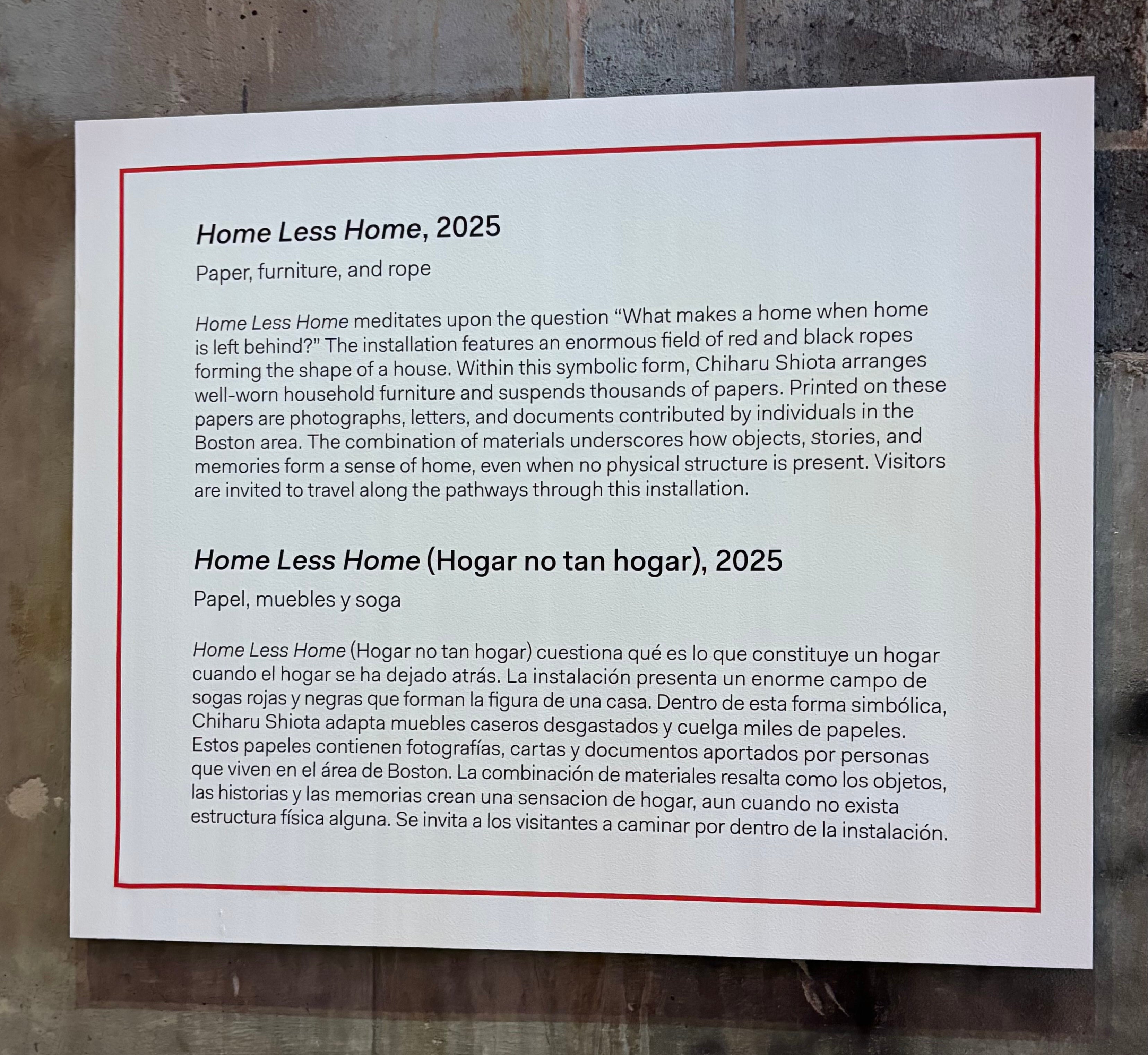 A large white label affixed to a concrete wall. The name of the work, the materials used, and a brief paragraph about the installation written in English; below, the name of the work, the materials used, and a brief paragraph about the installation written in Spanish. 
