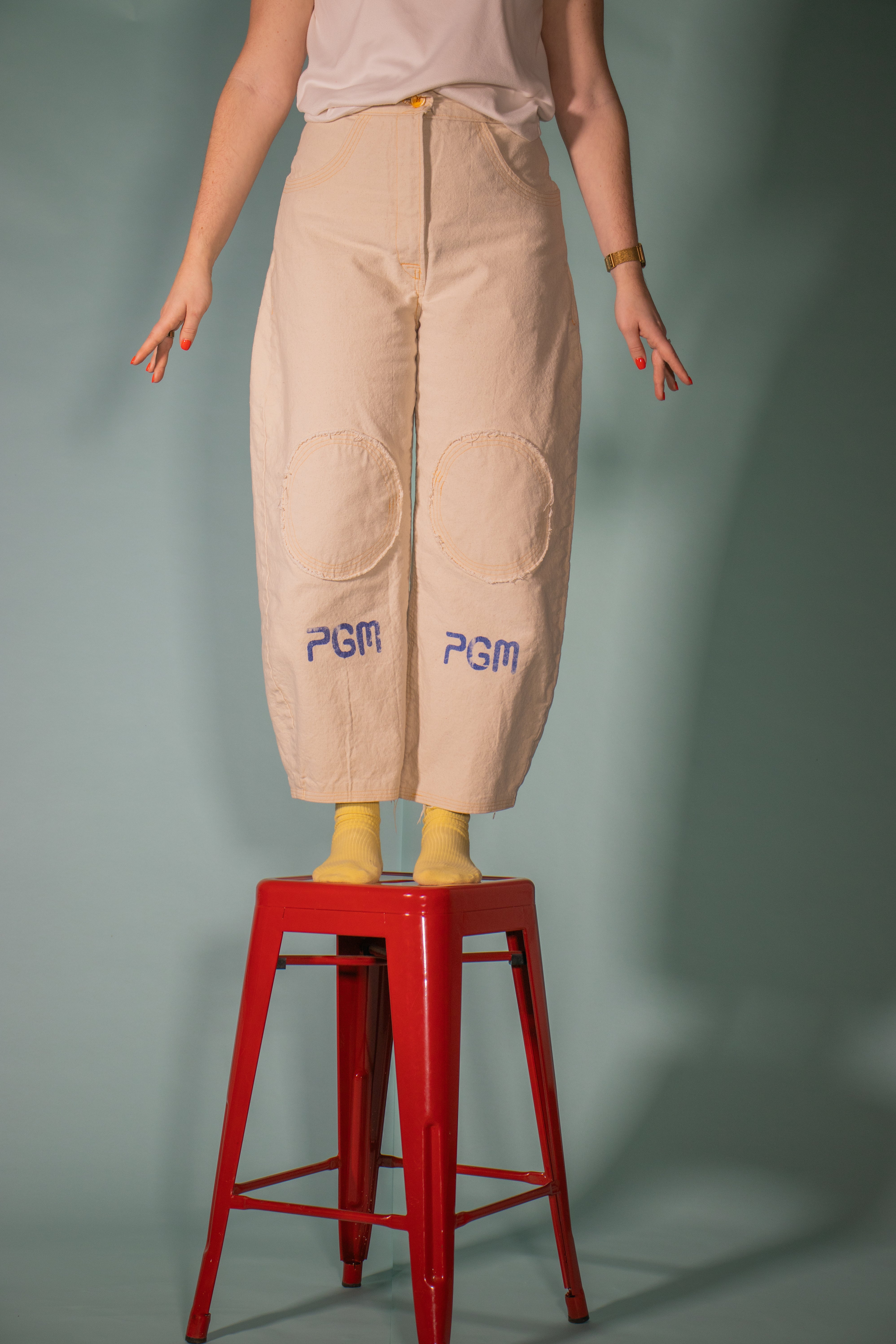 model standing on a red stool, showcasing canvas pants with her arms at her sides. you can't see her face, but you can see her yellow socks and the blue block print of the logo "PGM" under big circular knee pads finished with an orange button.