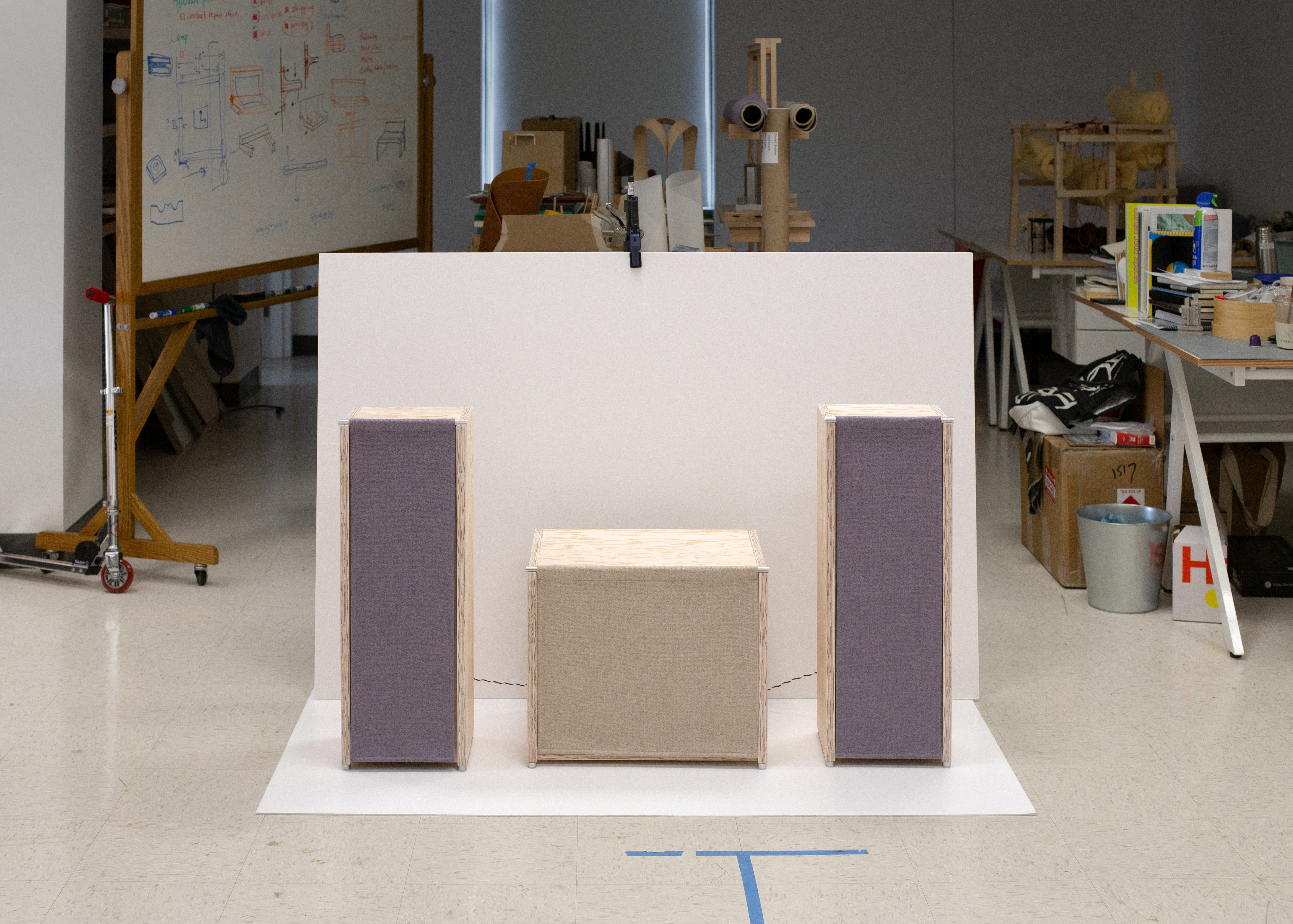 Two pale plywood speakers with hanging fabric fronts symmetrically flank a lower cabinet with a similar fabric front in a design studio, set against a large white backdrop with sketches, tools, and prototype furniture visible in the background.