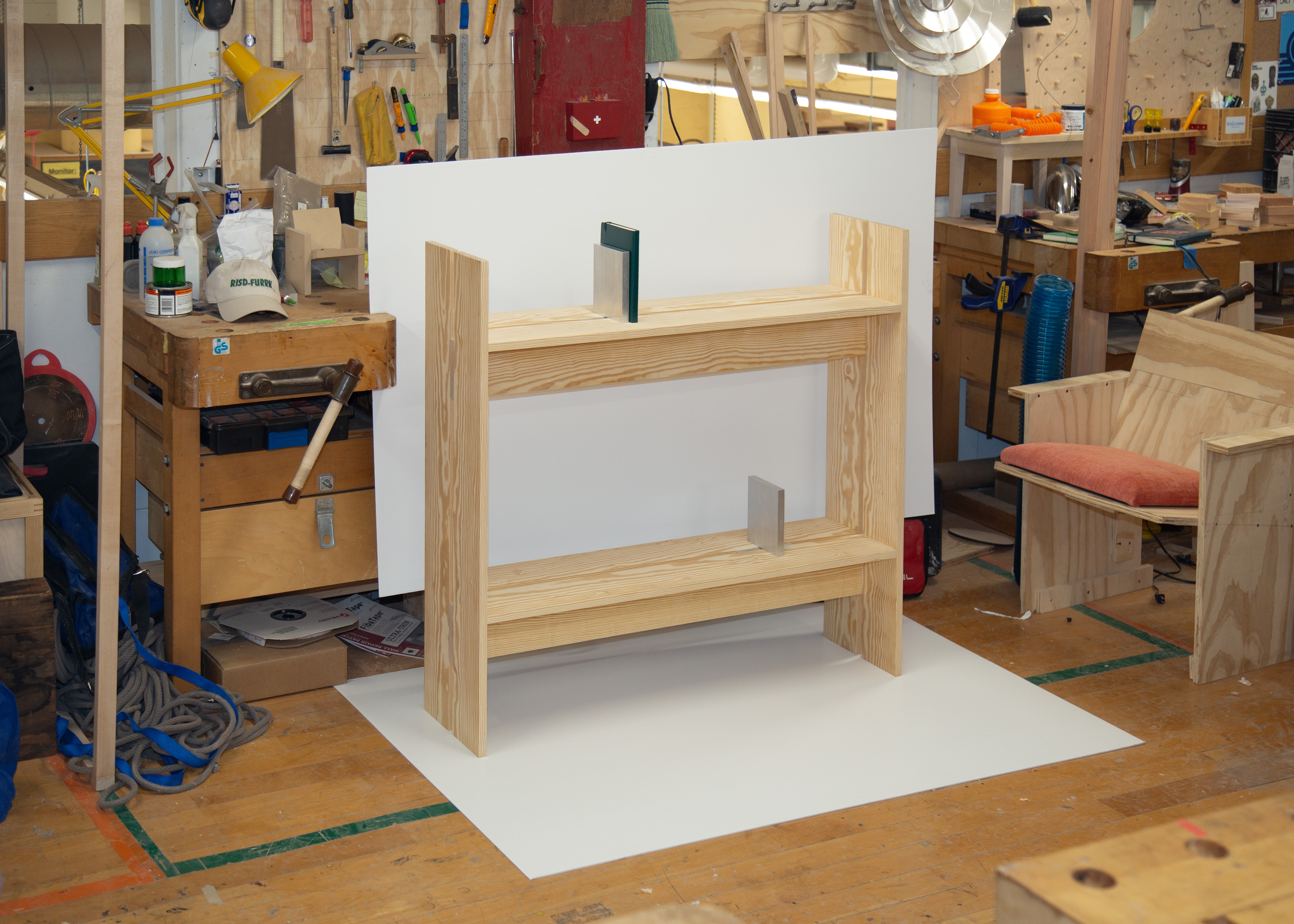 A pale wood shelving unit with integrated vertical book ends stands in a woodworking studio, displayed against a white backdrop amid workbenches, tools, and partially finished furniture prototypes.