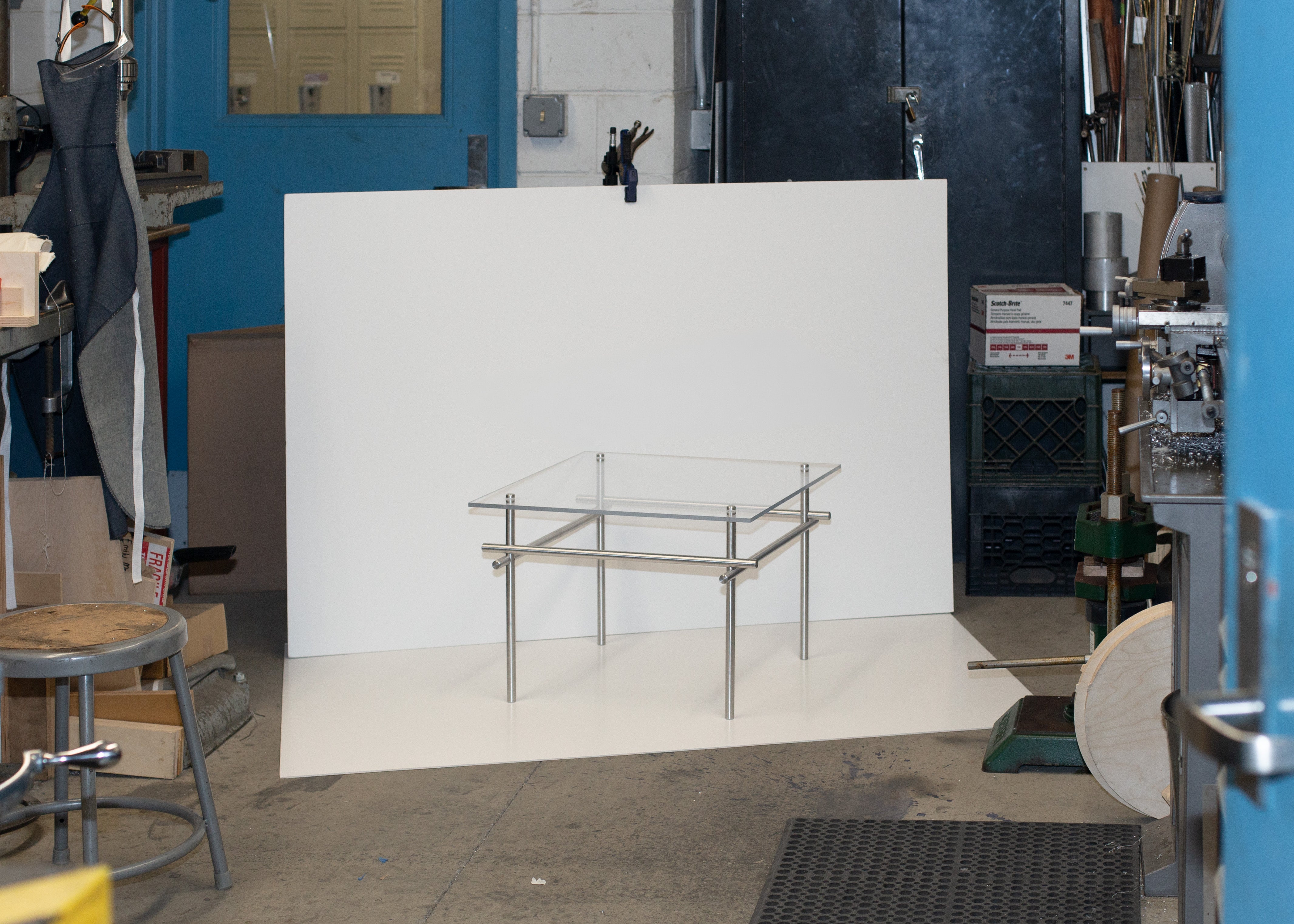 A small side table with a clear acrylic top and brushed metal rod frame is displayed against a white backdrop in a machine shop, surrounded by industrial tools, equipment, and workshop materials.