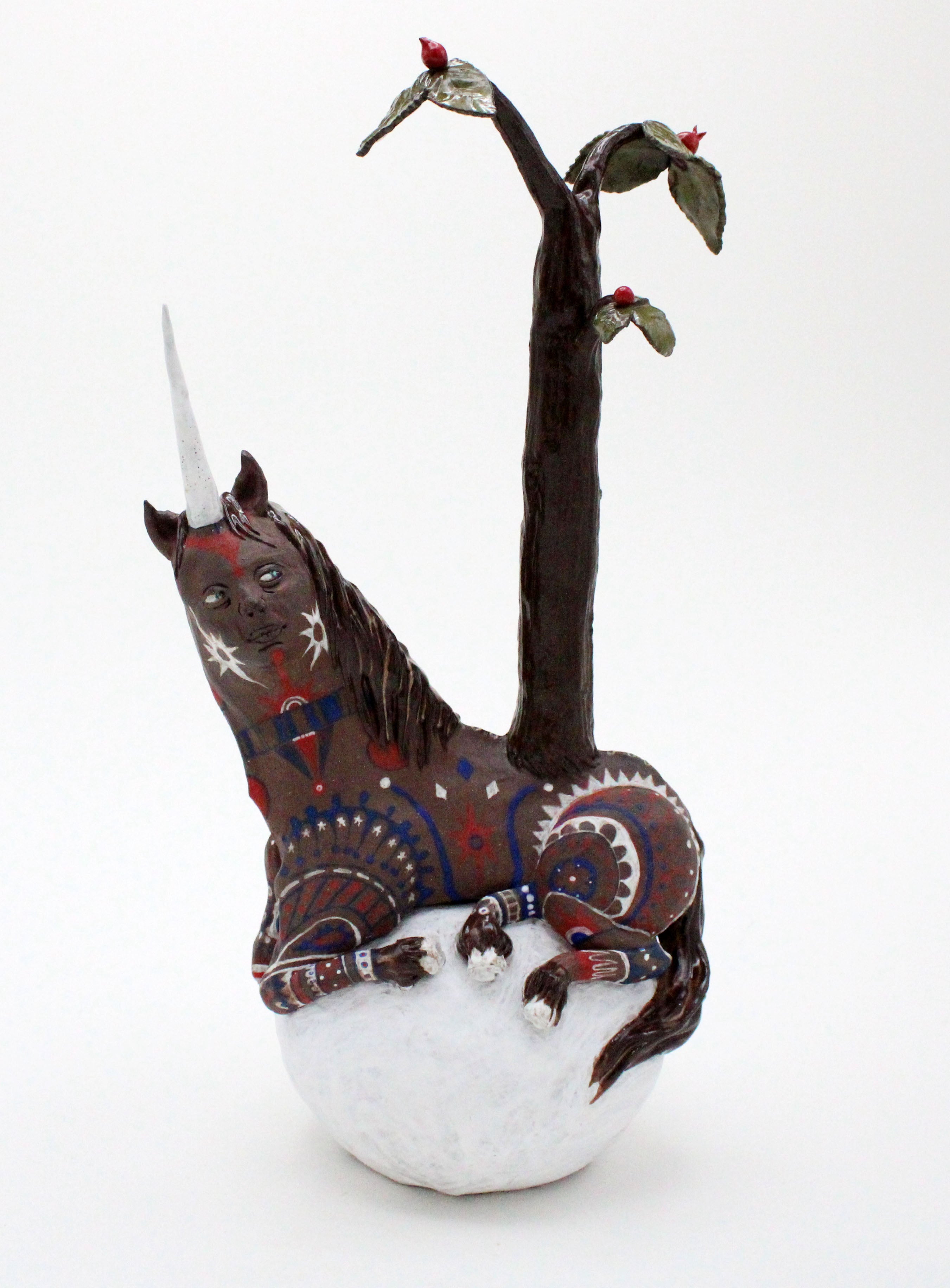 All ceramic: A brown unicorn on top of a white ball. It has the face of a woman and a tree growing through its back. It is covered in patterns inspired by Pictish warrior tattoos.