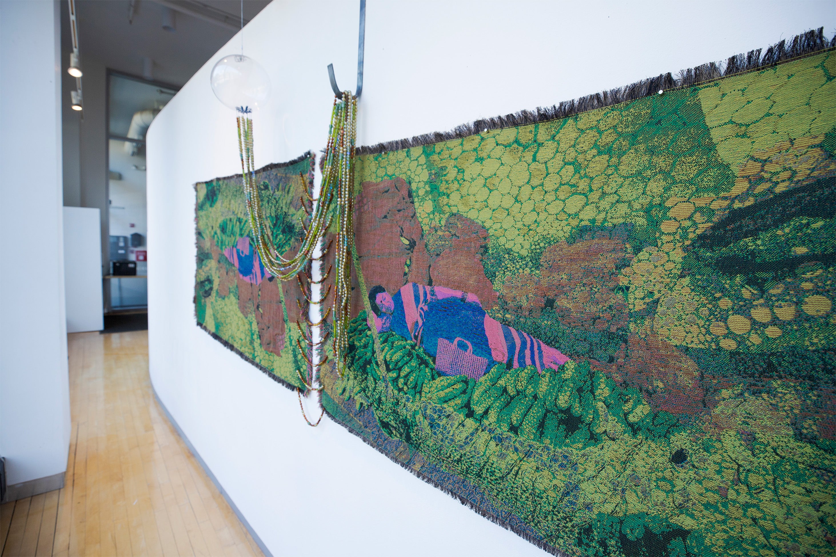 Jacquard-woven textile with lying figure among green forms, with draped glass beads connecting to a hanging glass orb