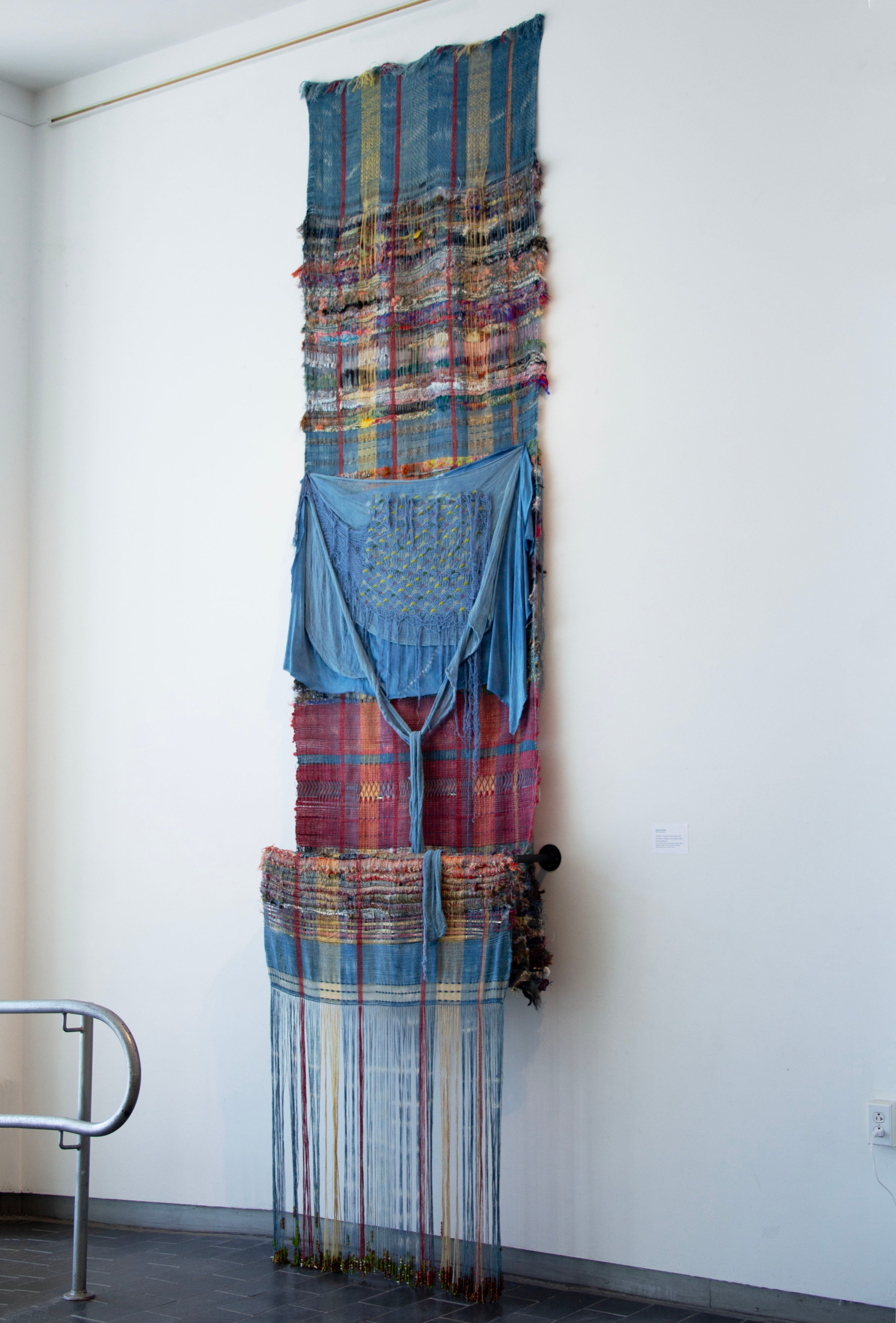 Vertical woven textile installation with layered blue and multicolored cloth, draped fabric, and long hanging thread beaded with glass beads