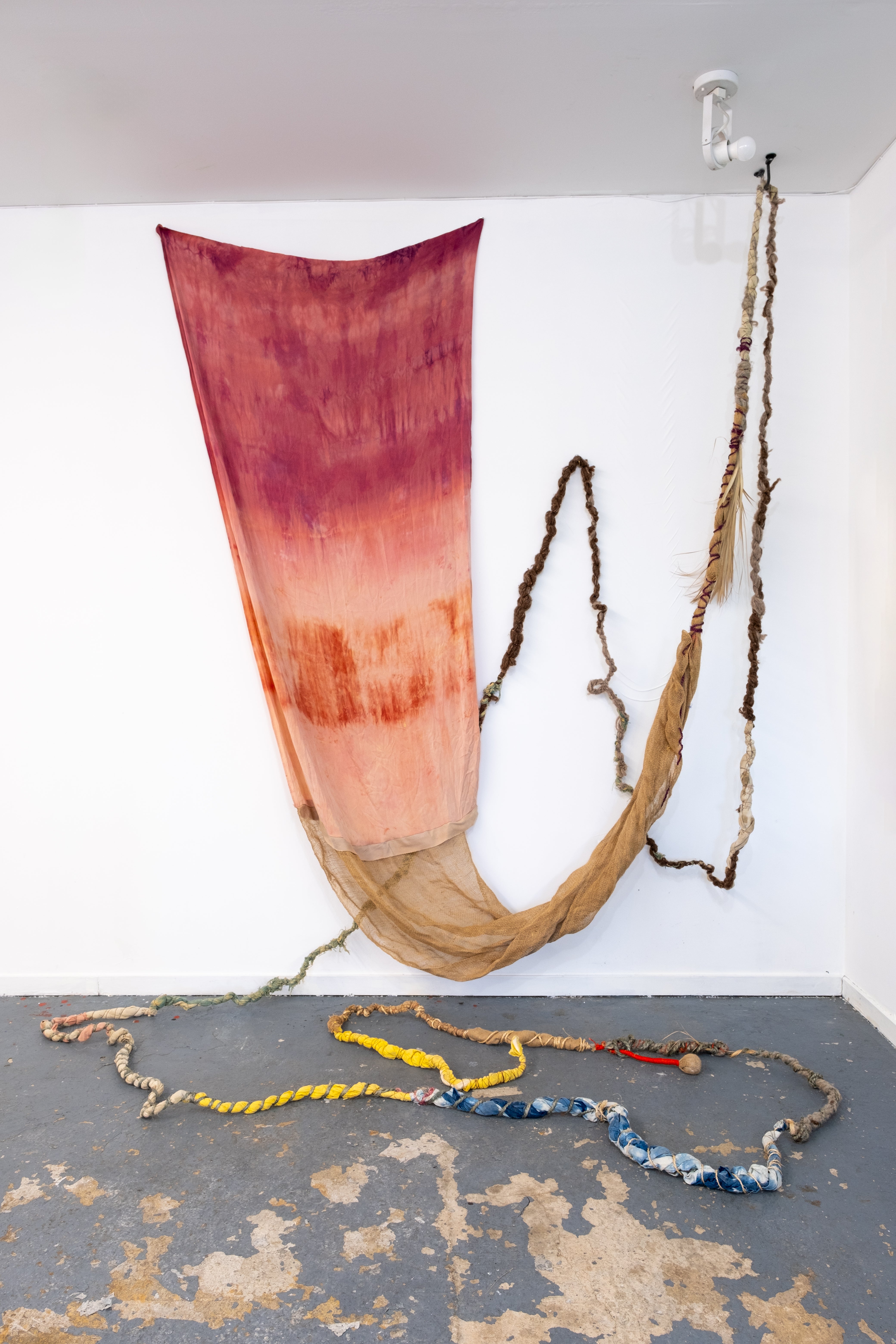 Hand-Natural Dyed textile in red and orange tones draped on wall, with rope-like fibers hanging and extending across the floor with small circle clay forms 