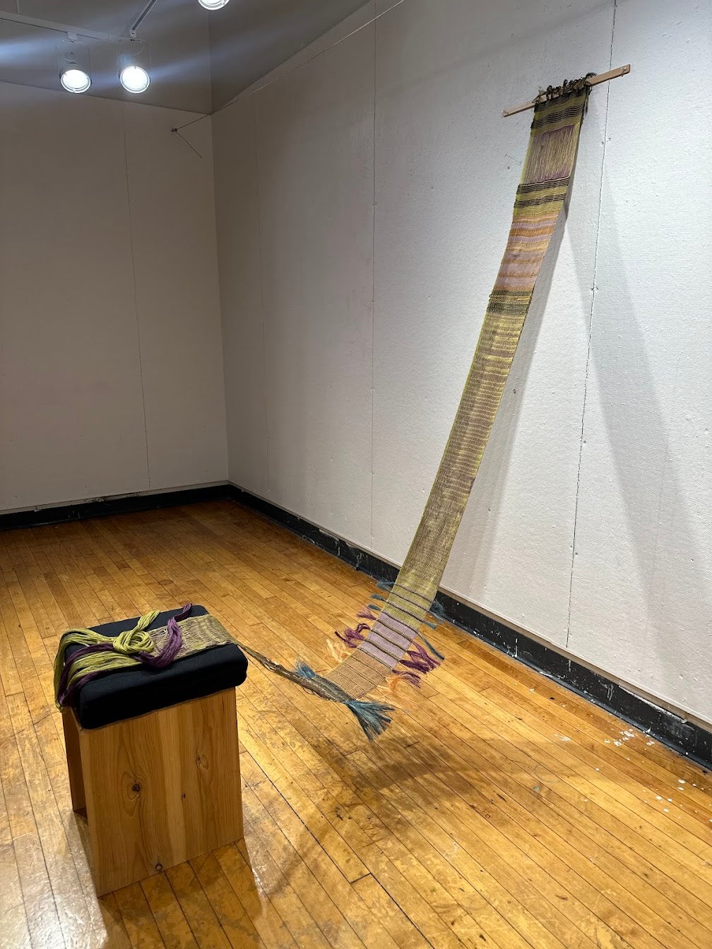 Narrow floor loom woven textile hanging from wall and trailing to floor, with striped patterns, draped onto a wooden pedestal with yarn