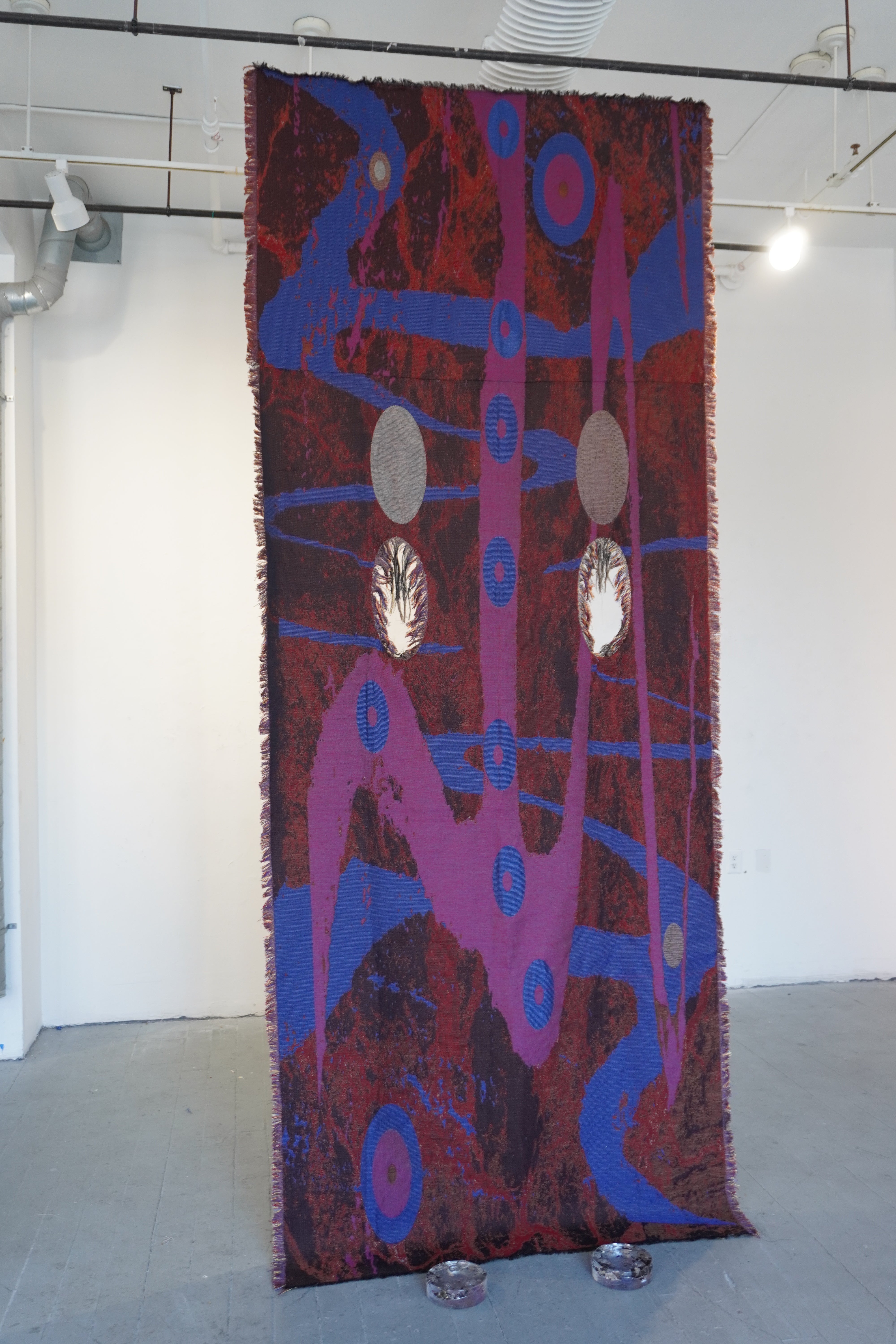 Tall hanging textile with red ground and blue abstract patterns, with reflective oval elements in a gallery space
