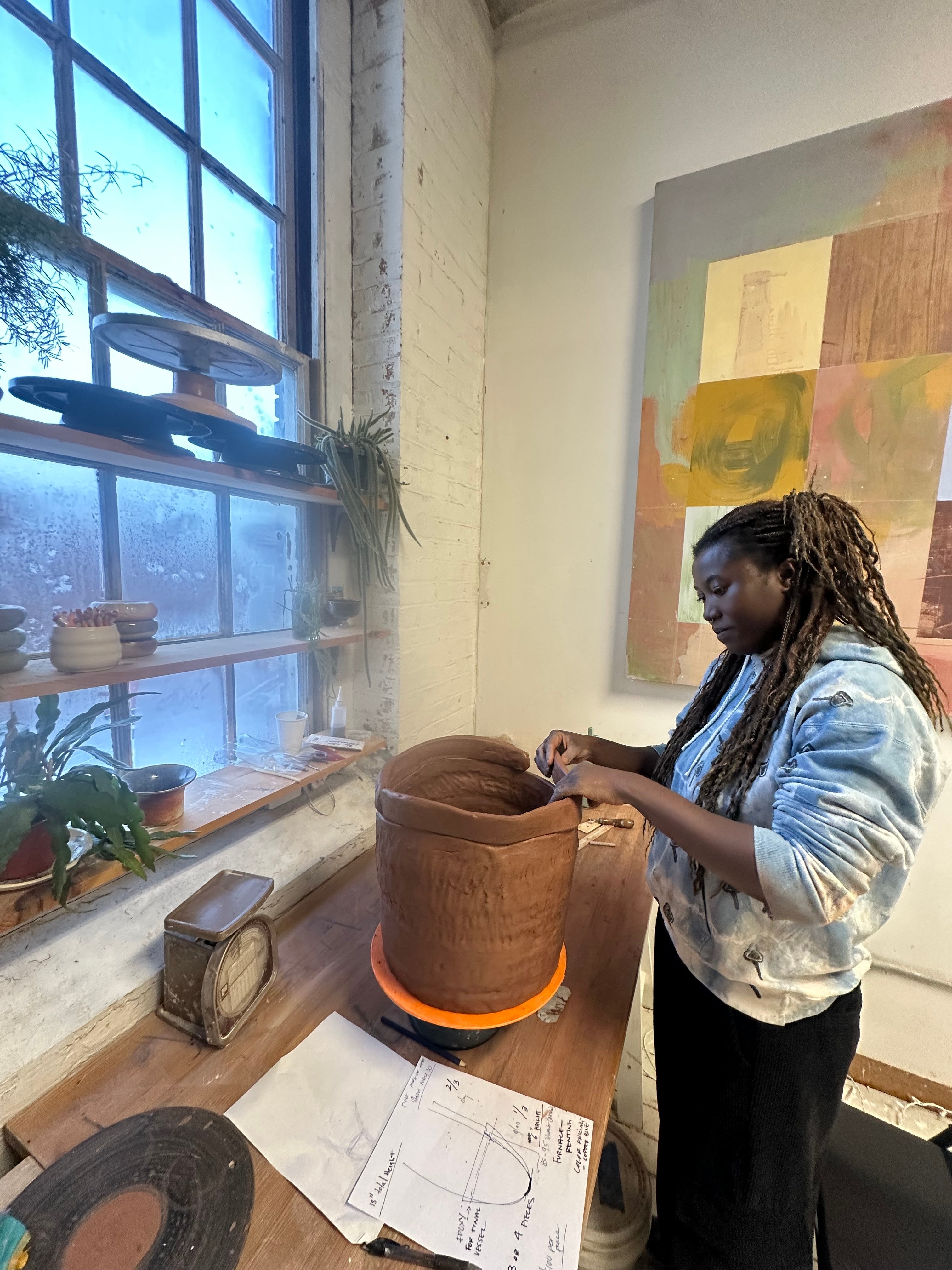Artist (Asmaa Amadou) working on a large clay vessel at a studio table by a window, with tools, plants, and sketches nearby