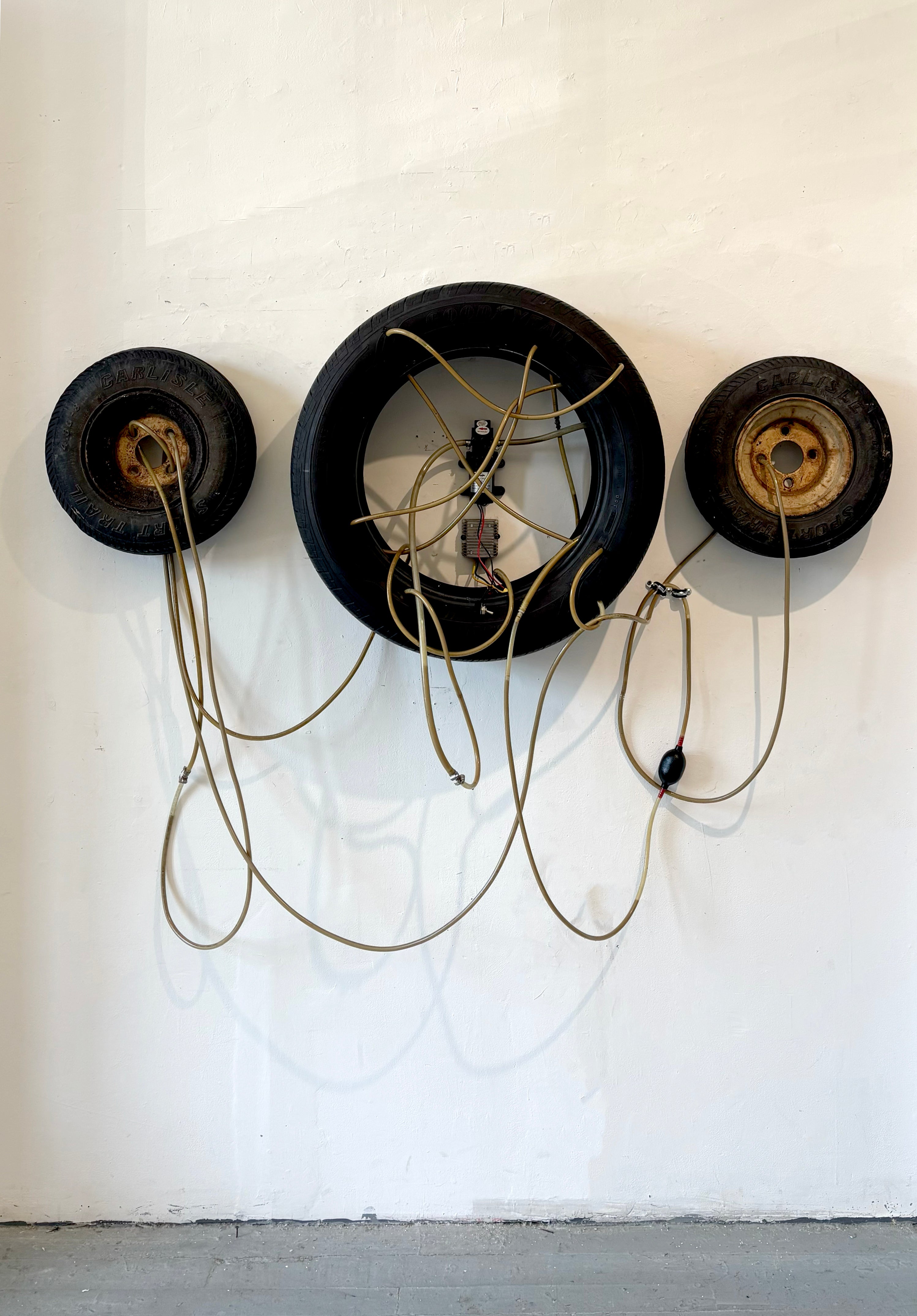 Three tires are assembled in a triptych format and are connected by a series of tubes and siphons. A motor is placed in the middle of the system and pumps liquid through allowing for some to leak out. 
