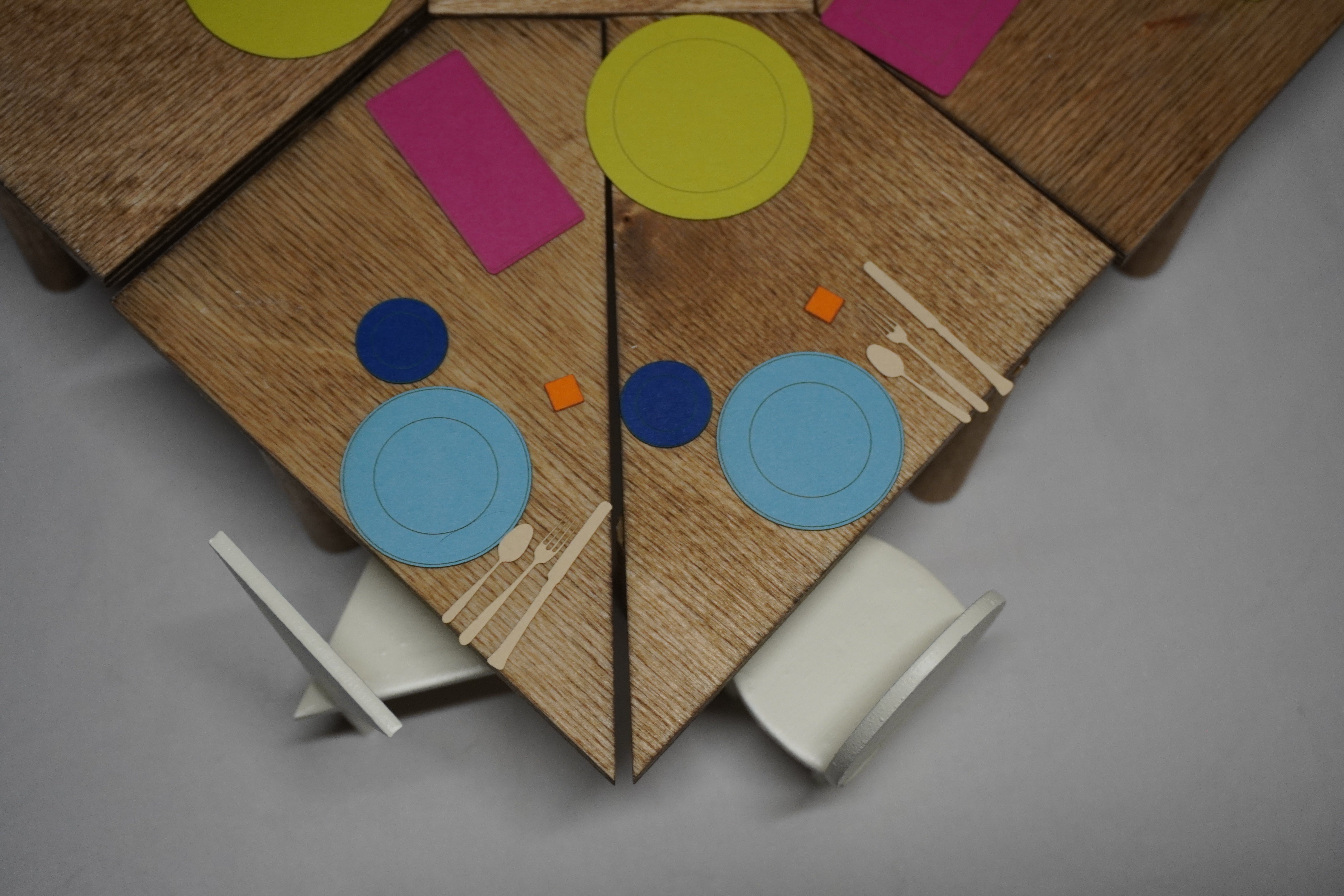 A corner of a table is shown from overhead view, where the pieces are broken in two, and  two different chairs sit adjacent to each other. Colorful kitchenware is scattered across the table surface. 