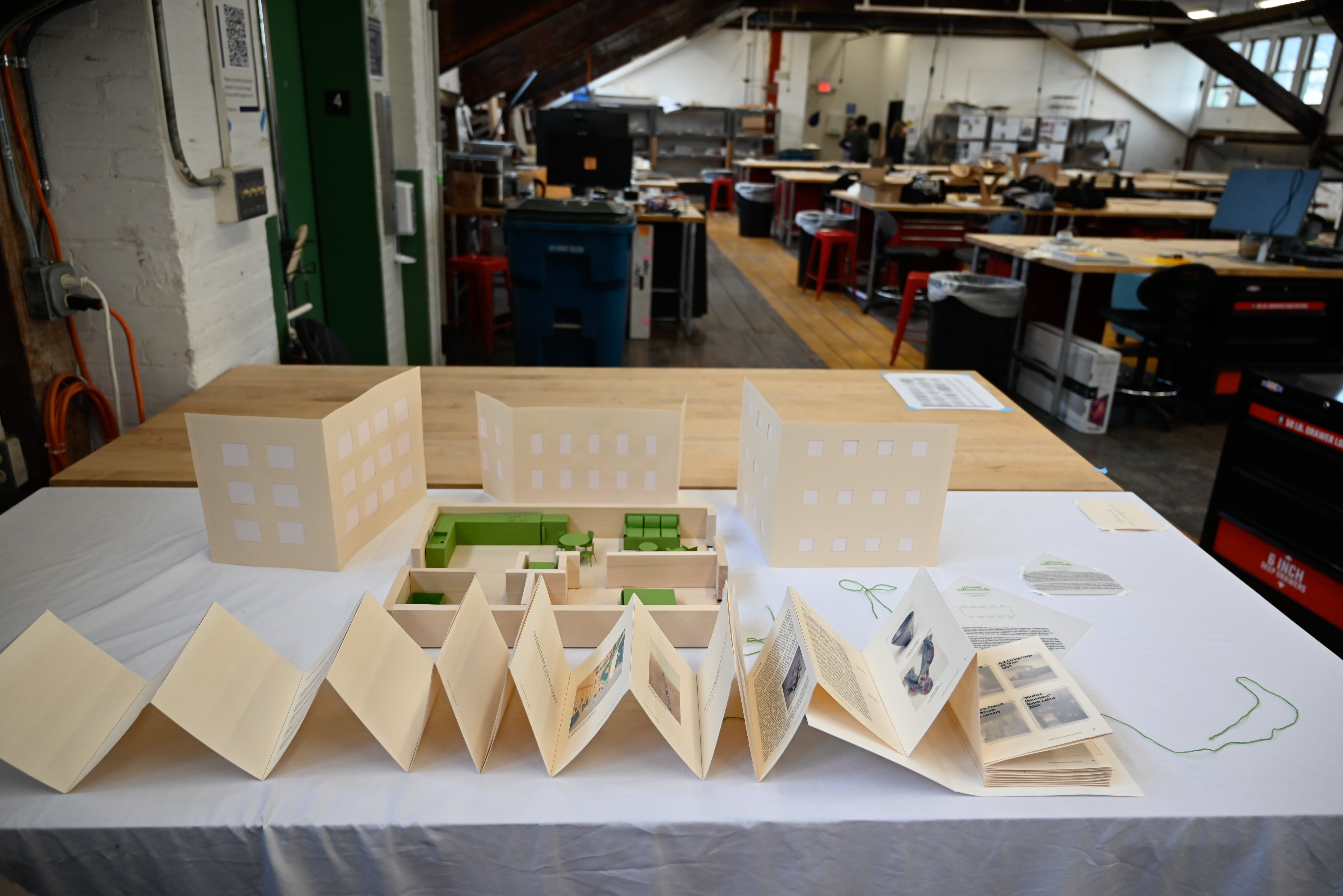 1/2" scale wooden model of one bedroom apartment and green paper furniture. Walls at 1/2 height. Apartment sits in front of paper building elevations. Accordion book spans the studio desk in front.
