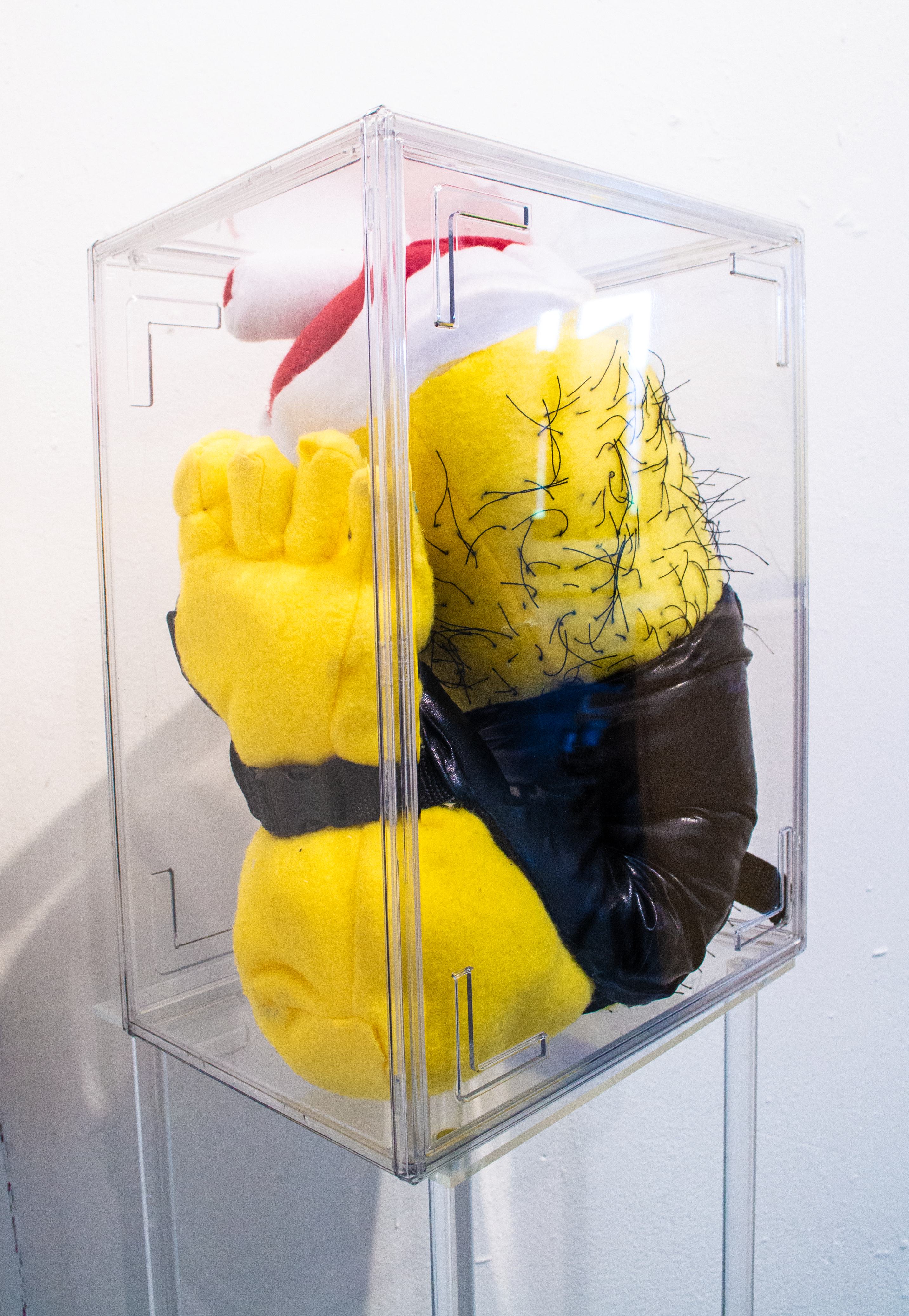 A clear acrylic case housing a plush, bright yellow severed foot. It is wearing a gladiator's boot. 