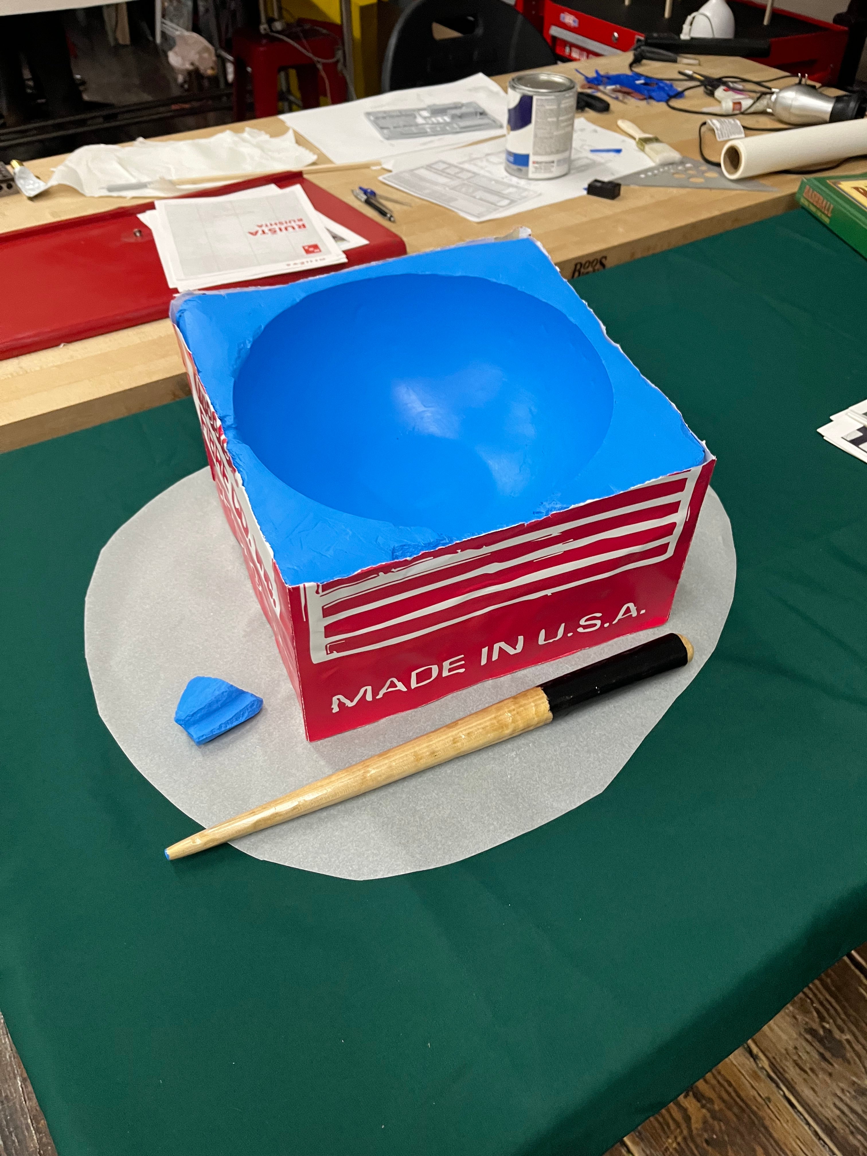 Process photo of an oversized blue pool cue chalk beside a miniature pool cue on a worktable. 