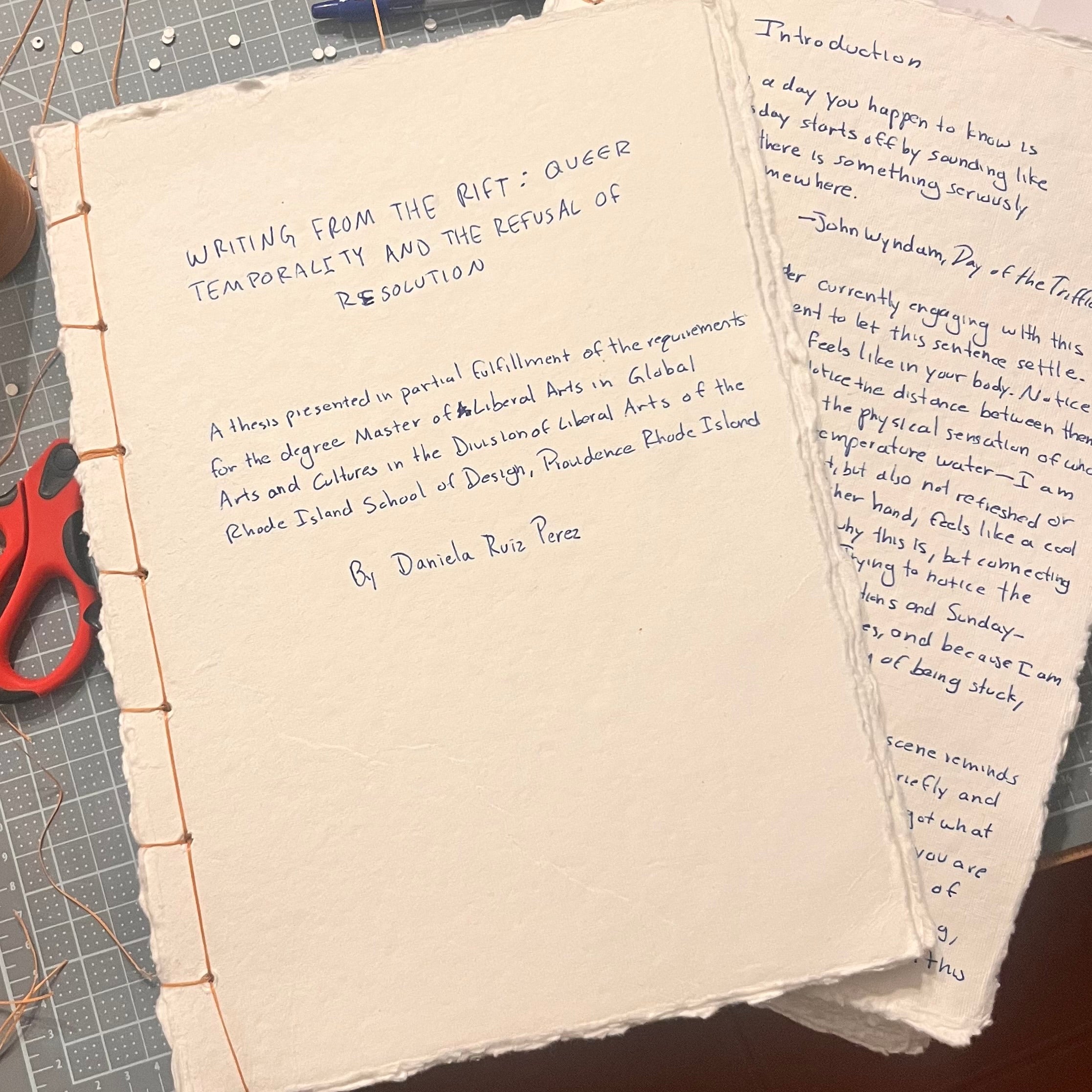 Hand-bound cream paper thesis manuscript on a cutting mat, stitched with copper thread along the left edge. Blue handwritten title reads “Writing From the Rift: Queer Temporality and the Refusal of Resolution.” Another handwritten page rests beside it.