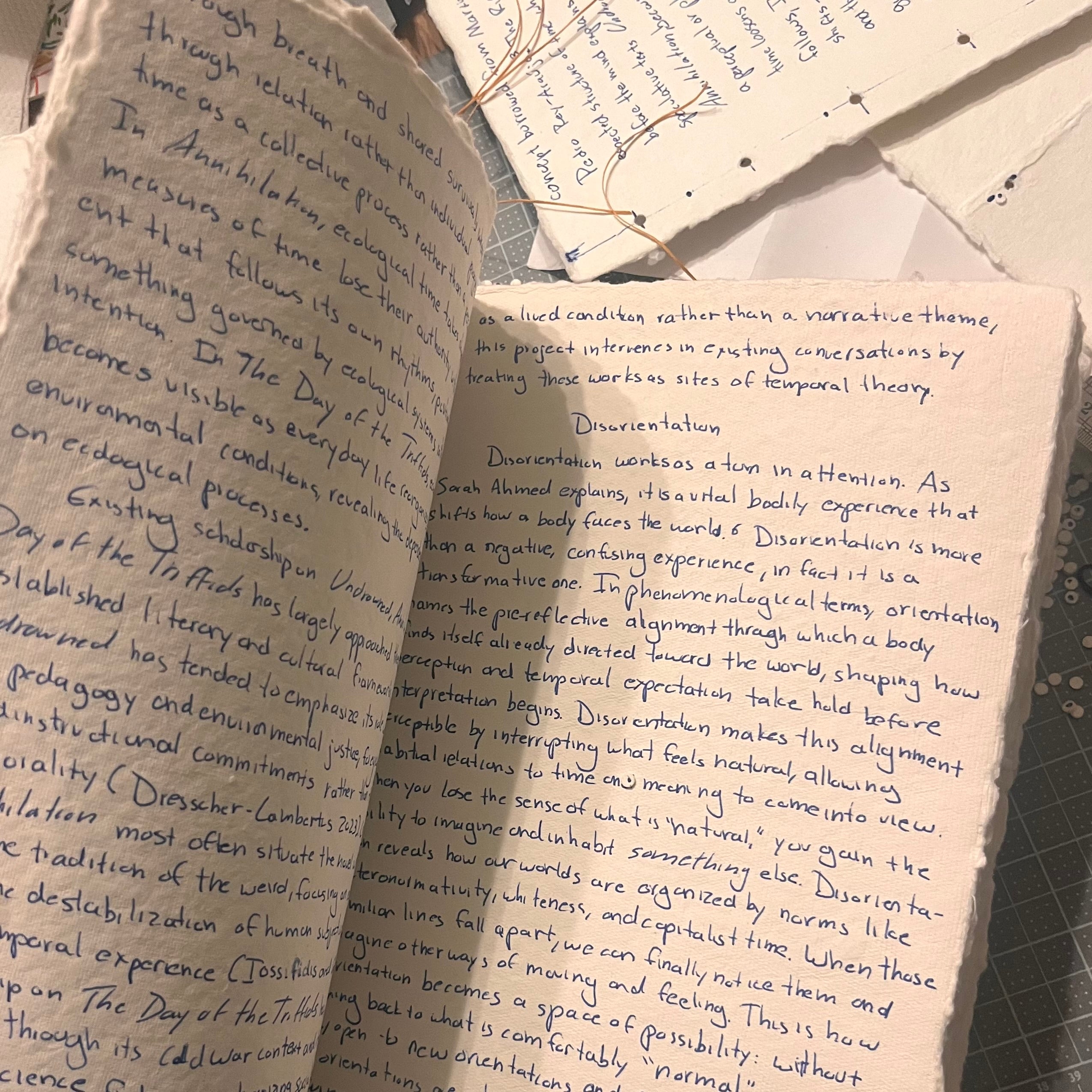 Open rough, cream paper thesis manuscript handwritten in blue ink sits on a cutting mat beside stitched pages and loose thread. Visible section titled “Disorientation” discusses Sarah Ahmed, phenomenology, and queer temporality.