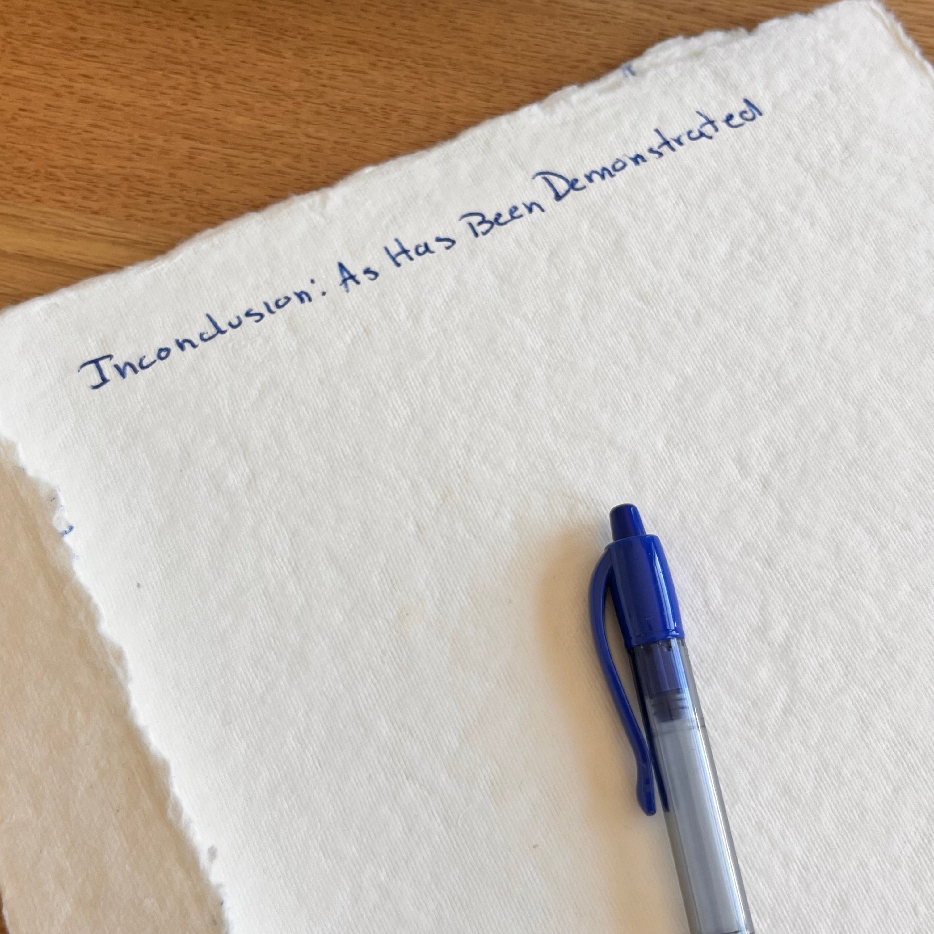 Cream paper with rough edges lies mostly blank on a wooden table beside a blue pen. Blue handwritten title at the top reads, “Inconclusion: As Has Been Demonstrated.