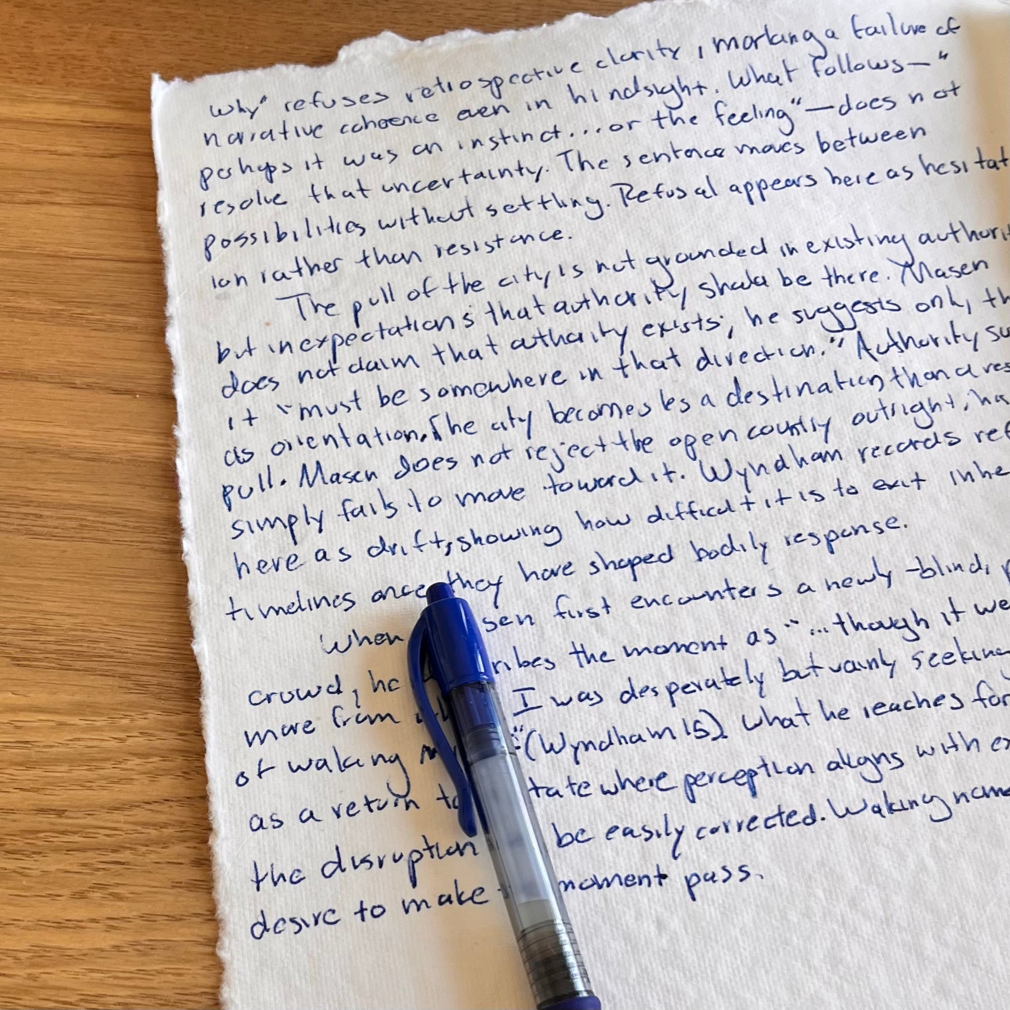 Deckled cream paper covered in blue handwritten thesis prose rests on a wooden table beside a blue pen. Text discusses refusal and uncertainty in relation to temporality and perception