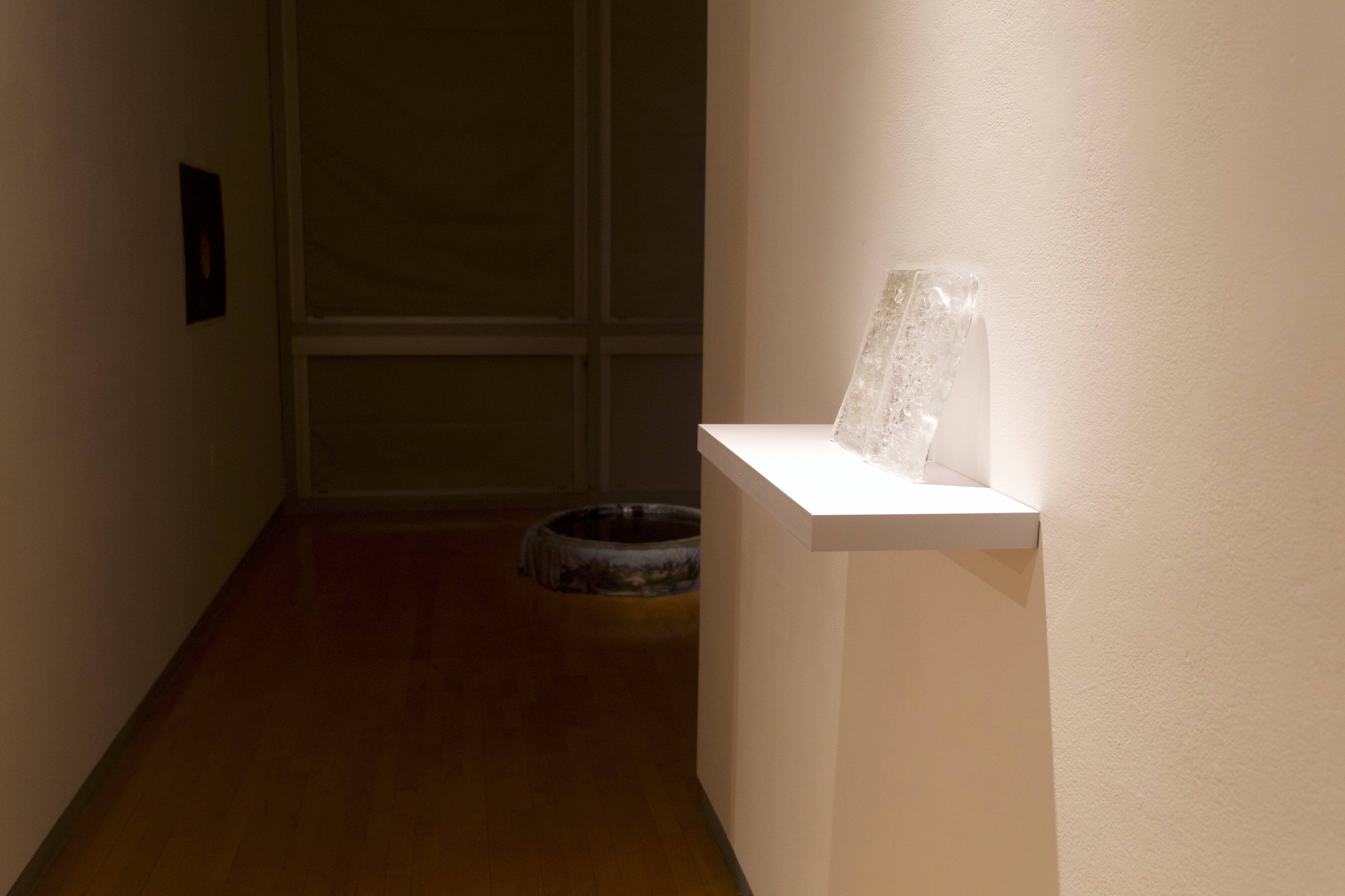 Dim installation view with translucent glass slabs on a white shelf and a dark water basin in the background.
