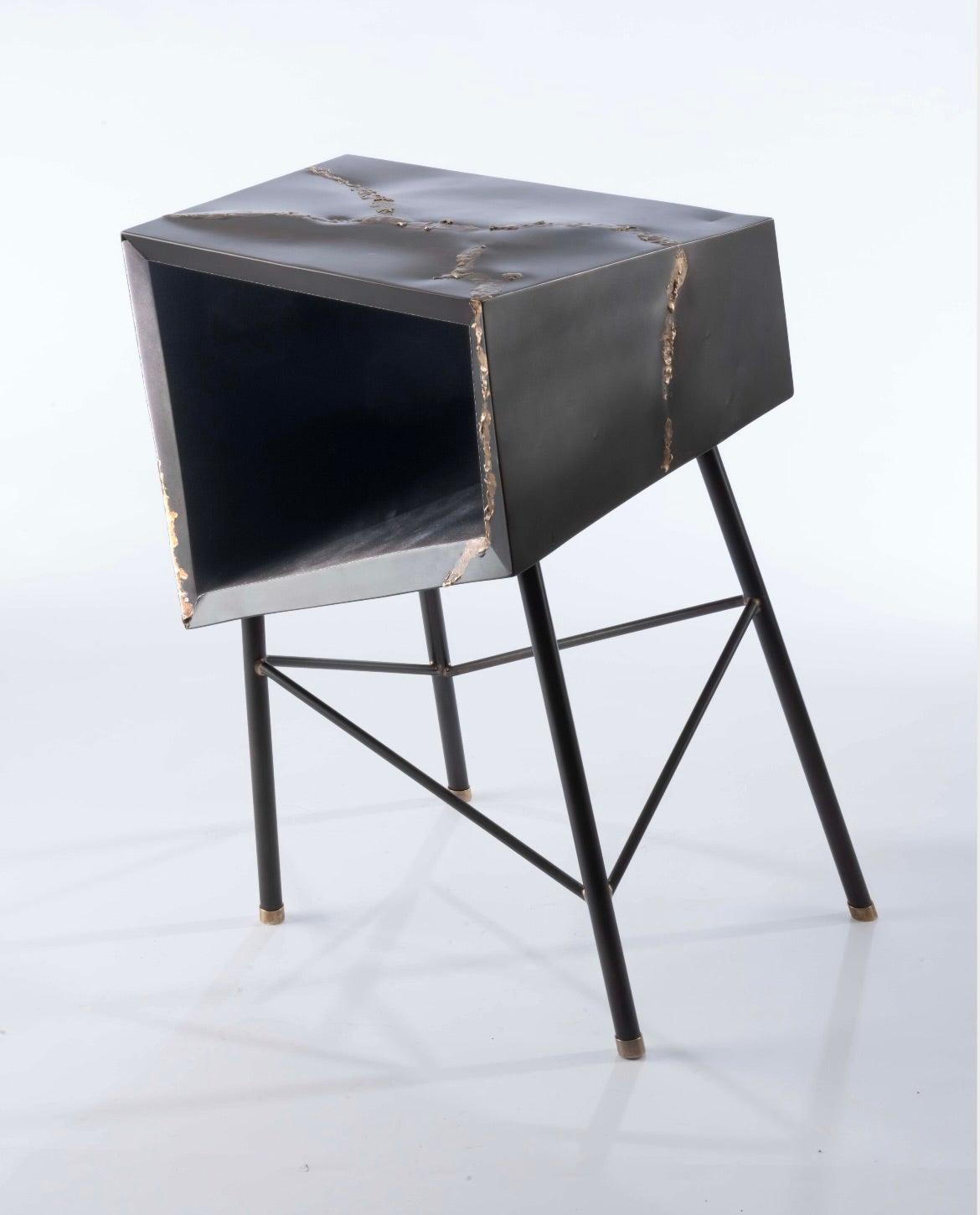 dark steel angular box sitting atop thin metal tubes arrange that seem askew, each connection has a gold fillet, there are brass feet and the corner shows melted bronze dripping down from the top surface