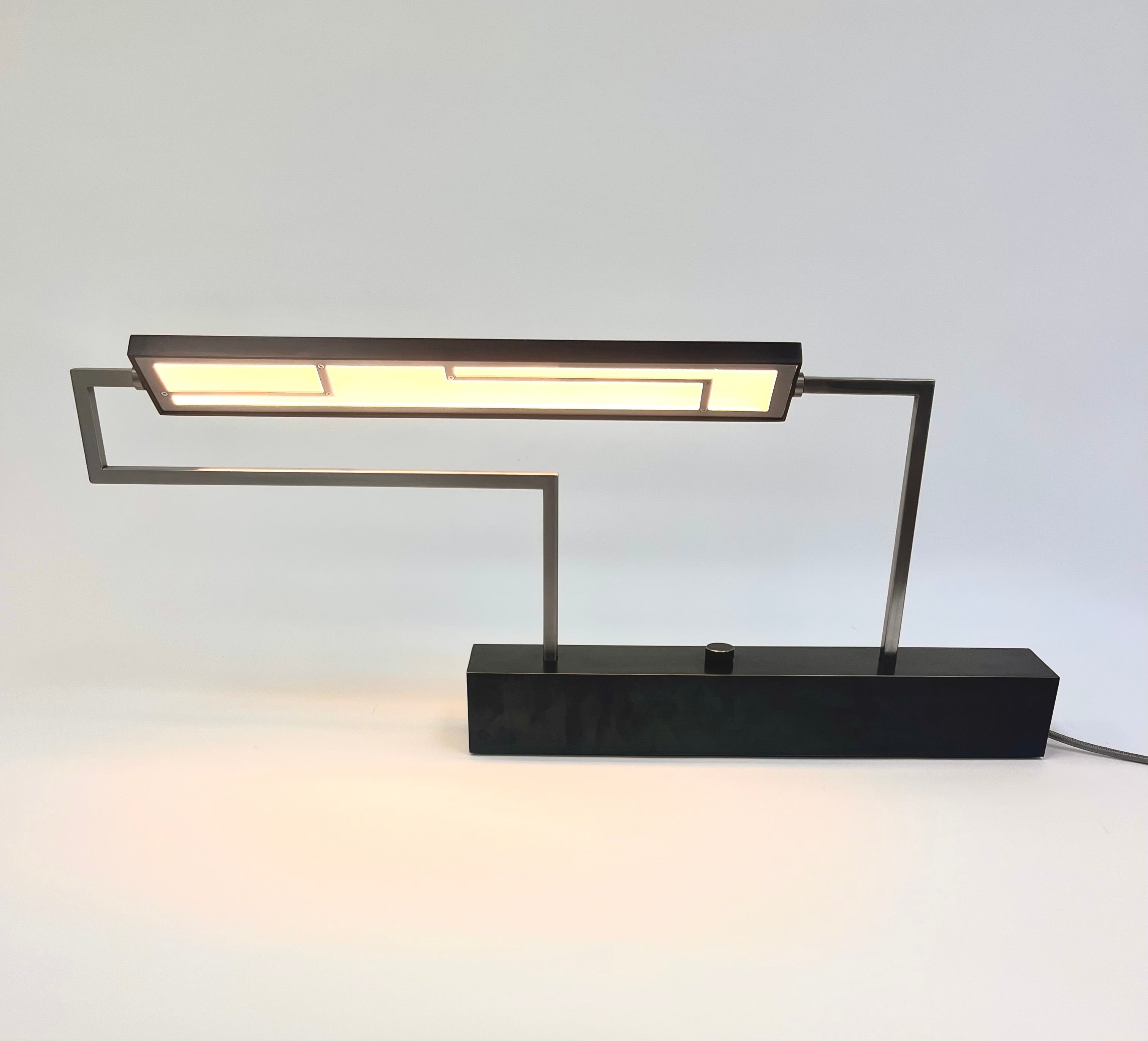 dark blue rectangular light with a patterned metal cover over diffuser, thin polished steel tube supports, larger dark blue rectangular base with a round steel knob for dimmer switch all corners highlighted by polished steel 