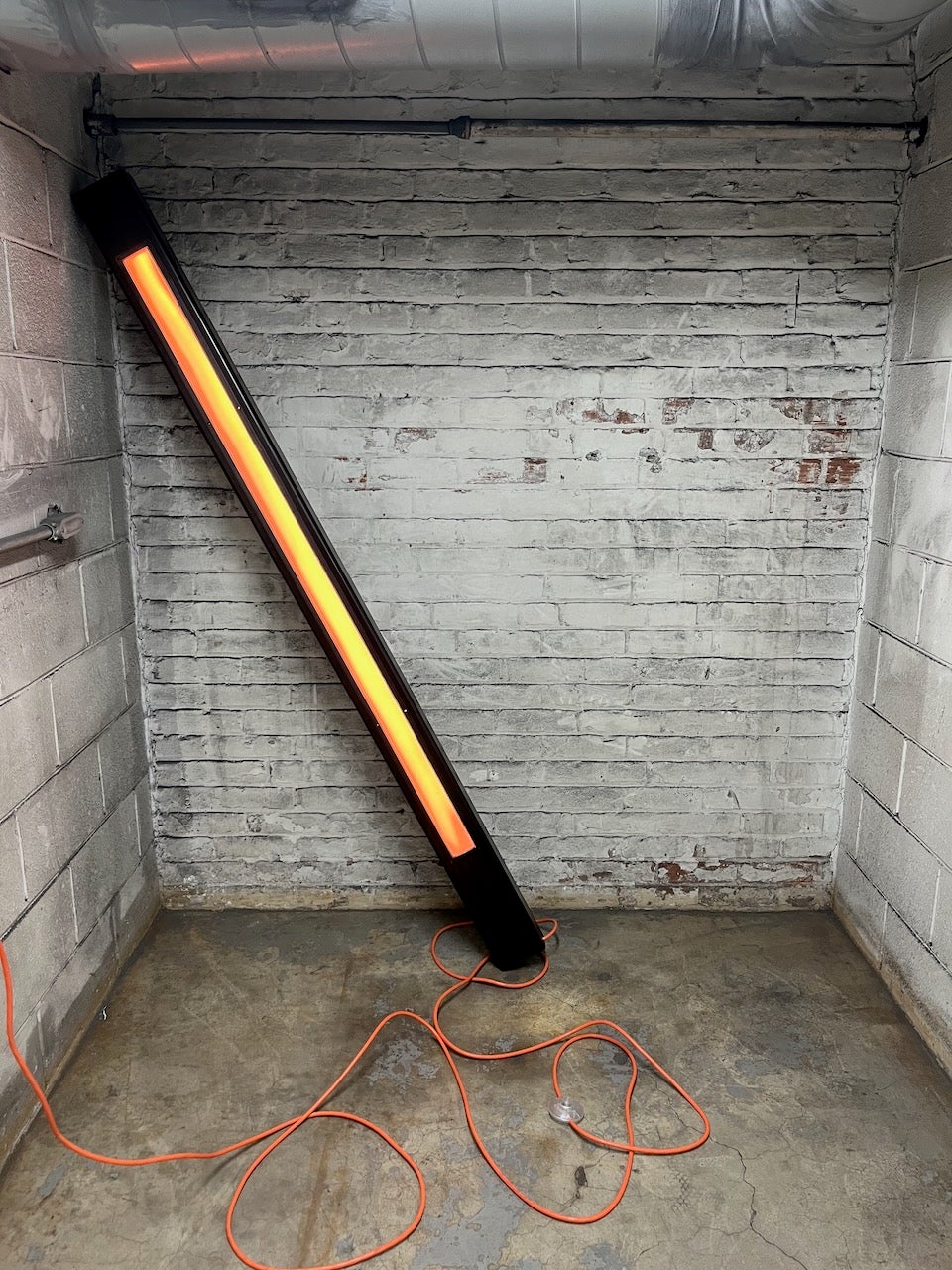 metal beam placed at very precarious angle in corner there is an orange beam of light on the front facing side of the large steel beam, and a jumble of extension cord at the base