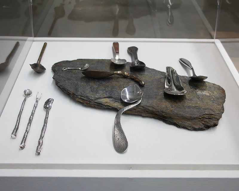 a series of spoons made from a variety of metals placed on and around a flat rock all arranged on a white pedestal enclosed in acrylic
